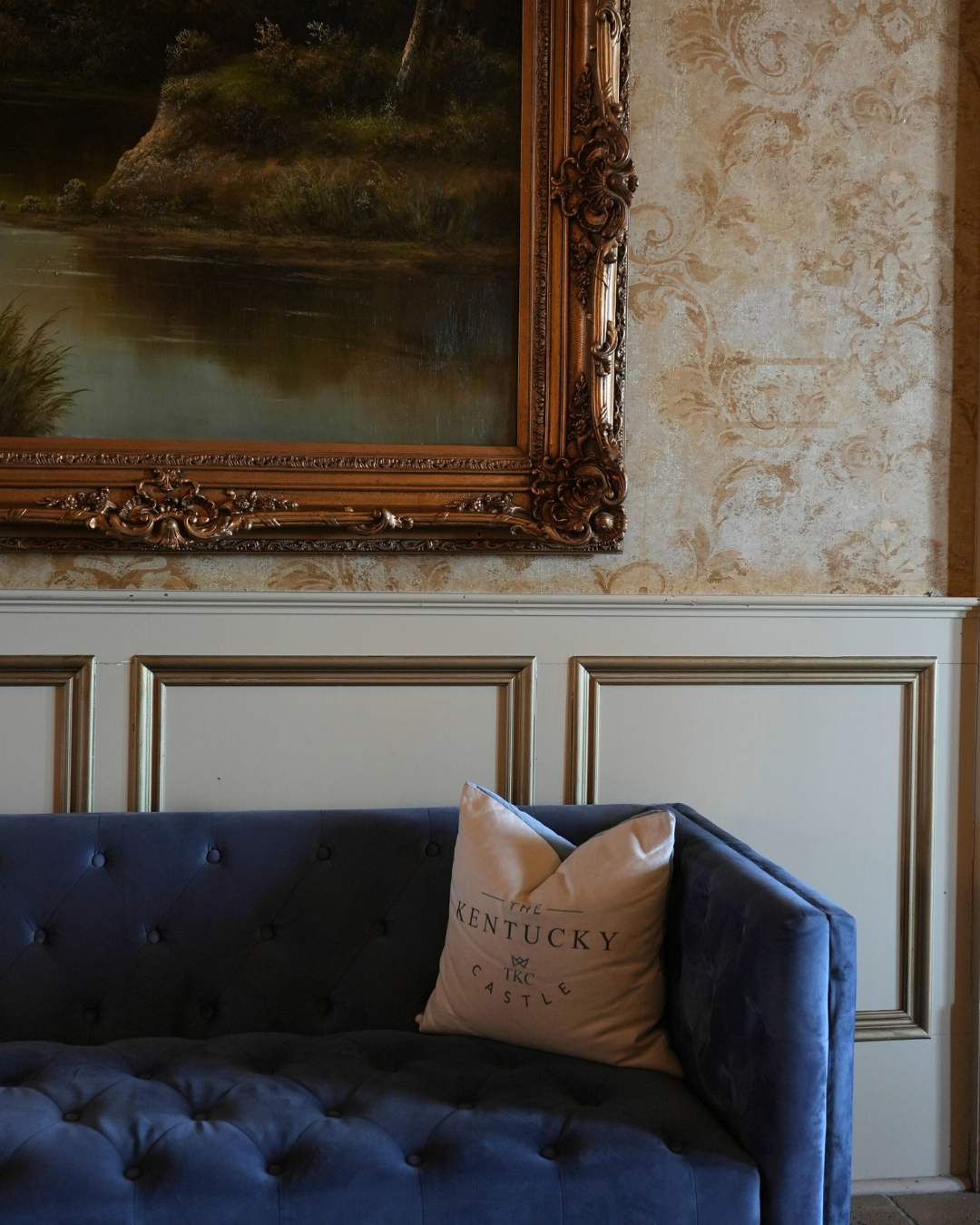 Blue velvet sofa with a Kentucky pillow under a framed landscape painting, against a patterned wall.