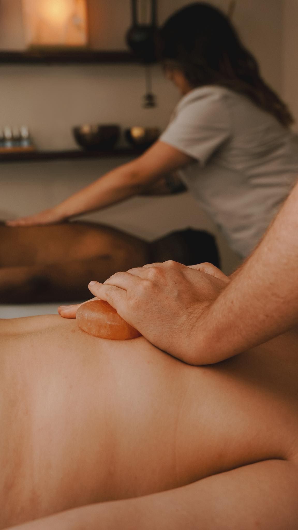 Hands performing massage with salt stone on a person's back, another person in the background.