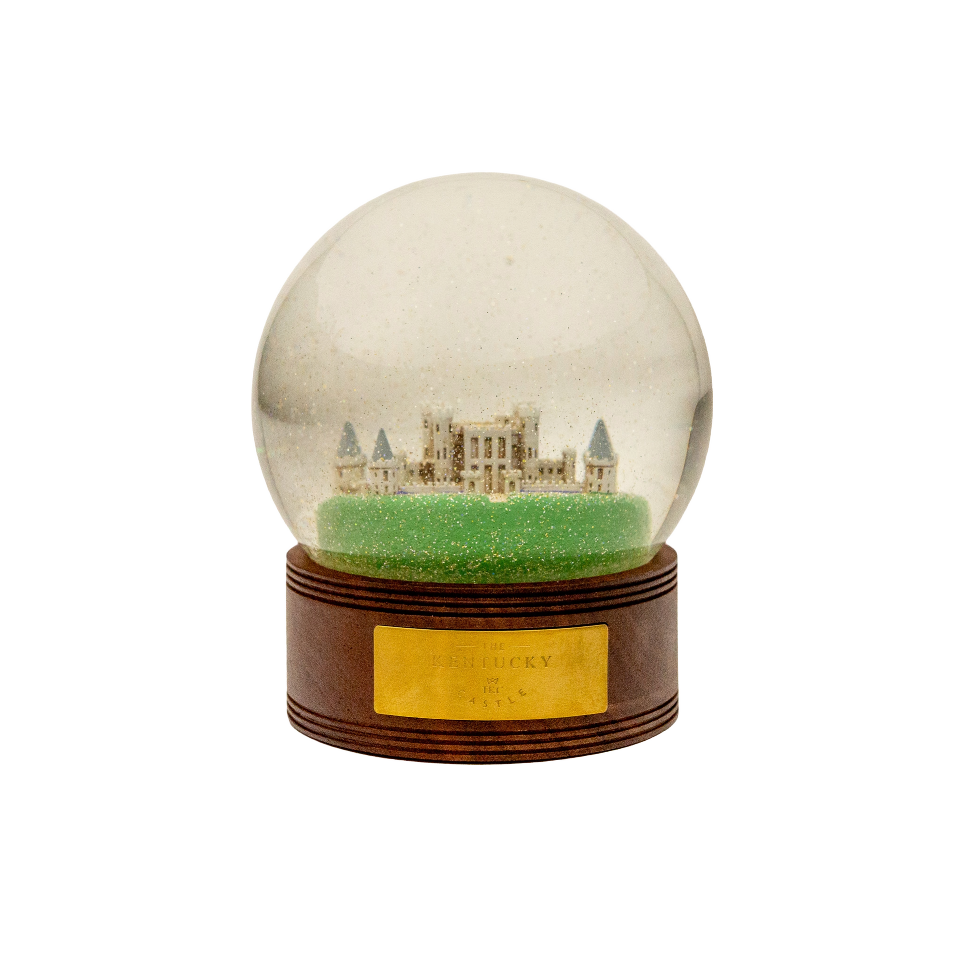Snow globe with a castle on a green base, atop a wooden stand, and a gold plaque.