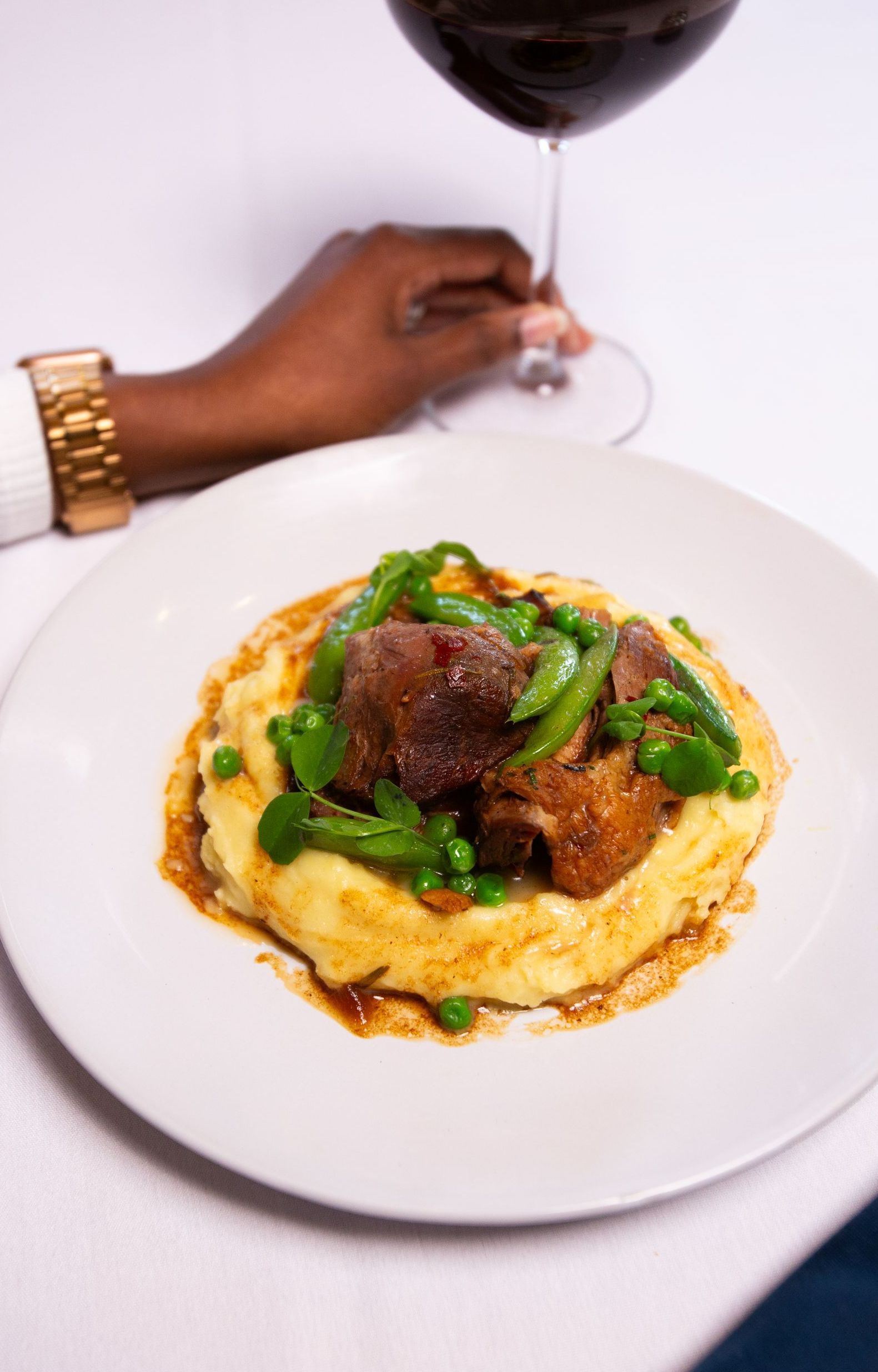 Braised meat with peas on mashed potatoes, a hand holding red wine glass, white tablecloth.