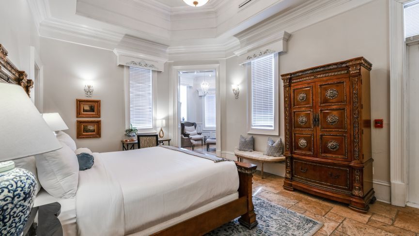 Elegant bedroom with ornate wooden armoire, bed, windows, and access to a sitting area.
