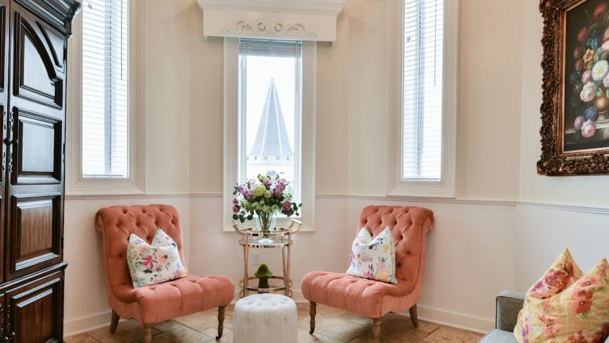 Two coral armchairs flank a small table with flowers, in a room with a window and floral painting.