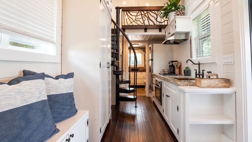 Interior of a tiny house with a kitchen, stairs, and seating area; white walls, wood floors, and blue accents.
