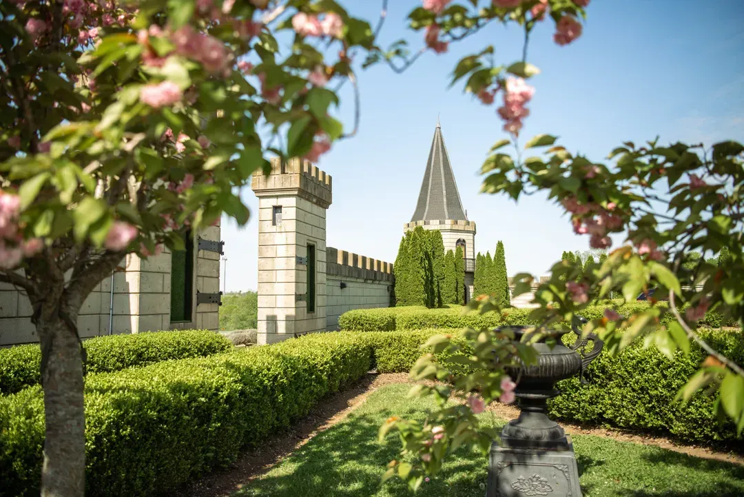 Stone castle with a pointed spire, surrounded by hedges and flowering trees on a sunny day.