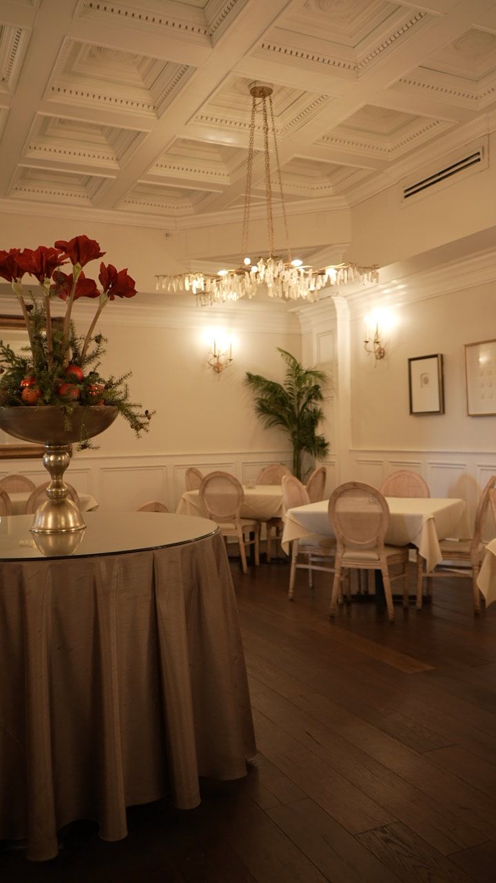 Elegant restaurant interior with round table, chairs, chandelier, and flowers. White walls, dark wood floor.