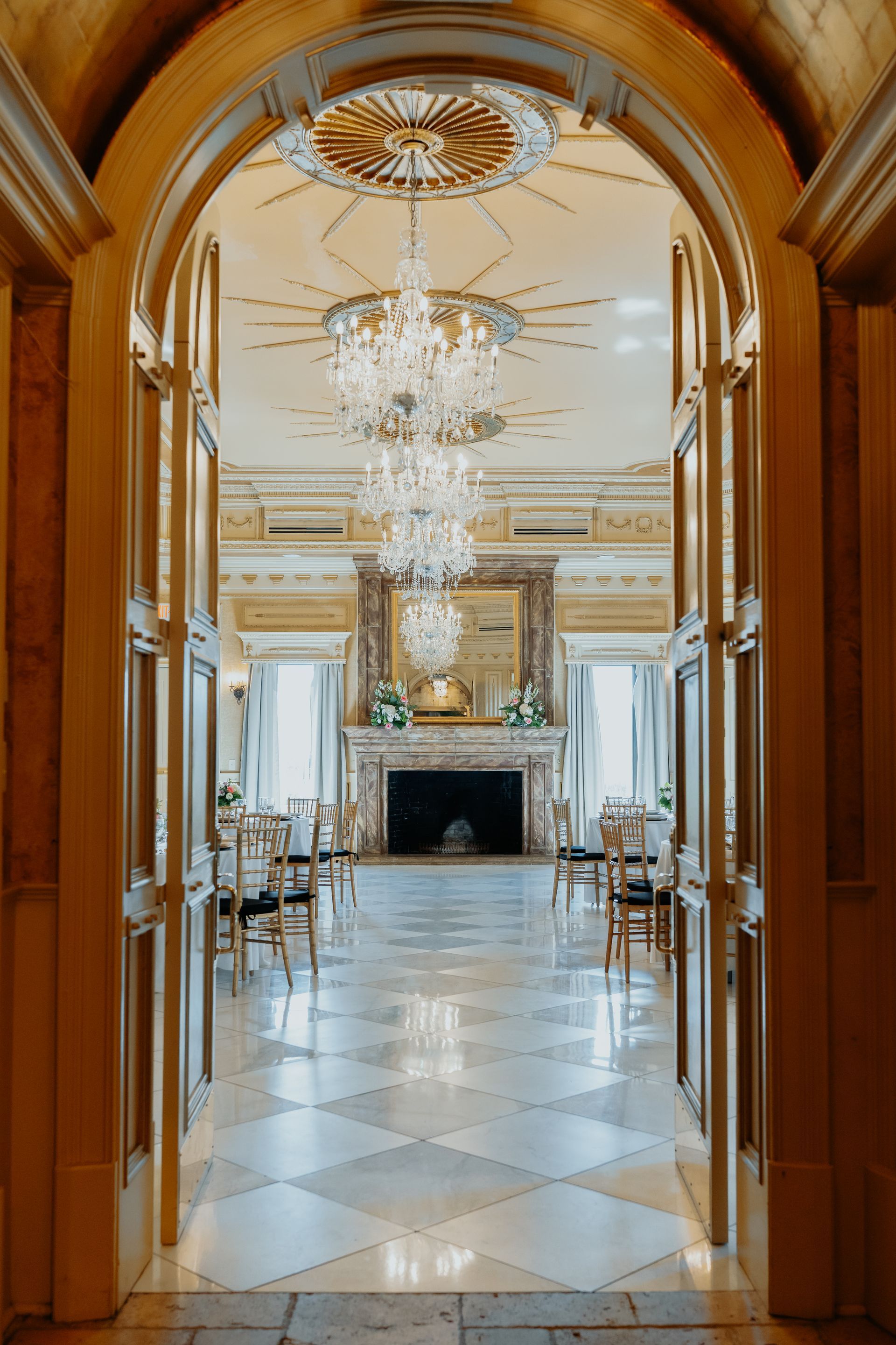 Golden archway frames an opulent ballroom with chandeliers, marble floor, fireplace, and chairs set up.