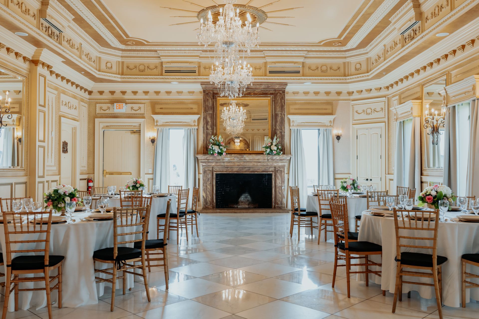 Elegant ballroom set for an event with tables, chairs, ornate gold and white decor, and a chandelier.