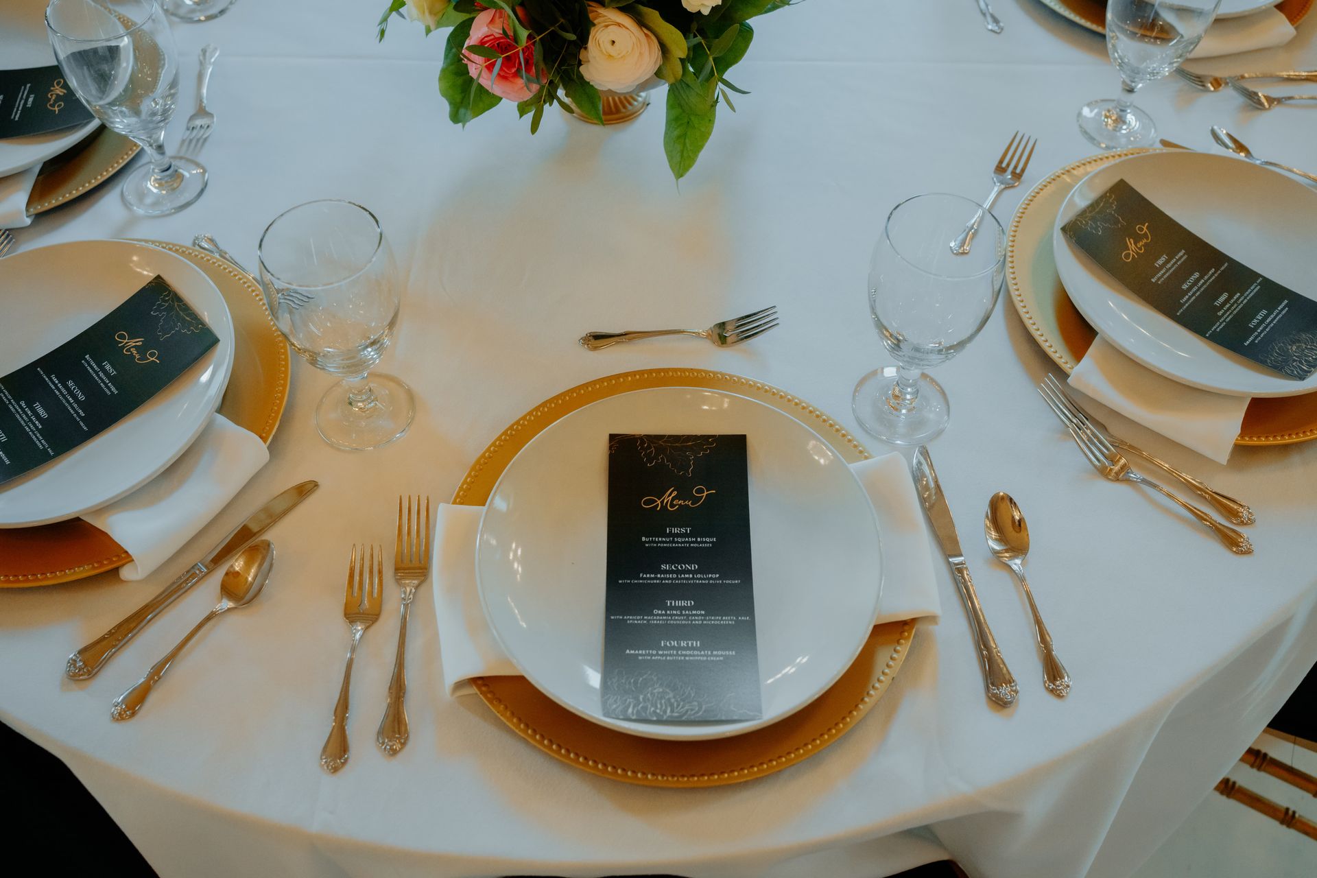 Elegant table setting with gold chargers, white plates, silverware, and a menu, centered around a floral arrangement.
