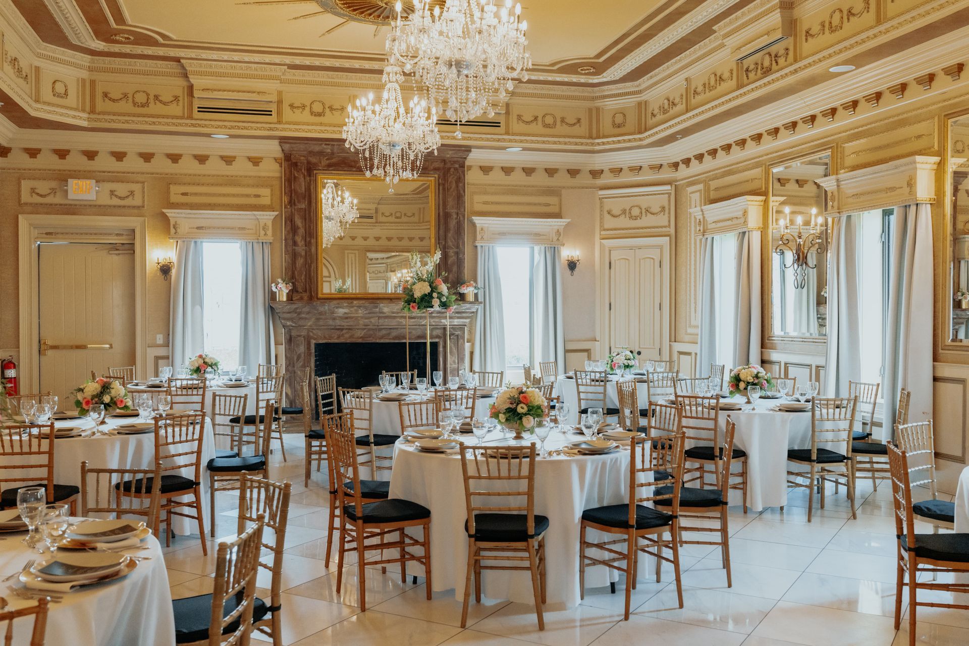 Elegant banquet hall with round tables set for a formal event; chandeliers, fireplace, gold accents.