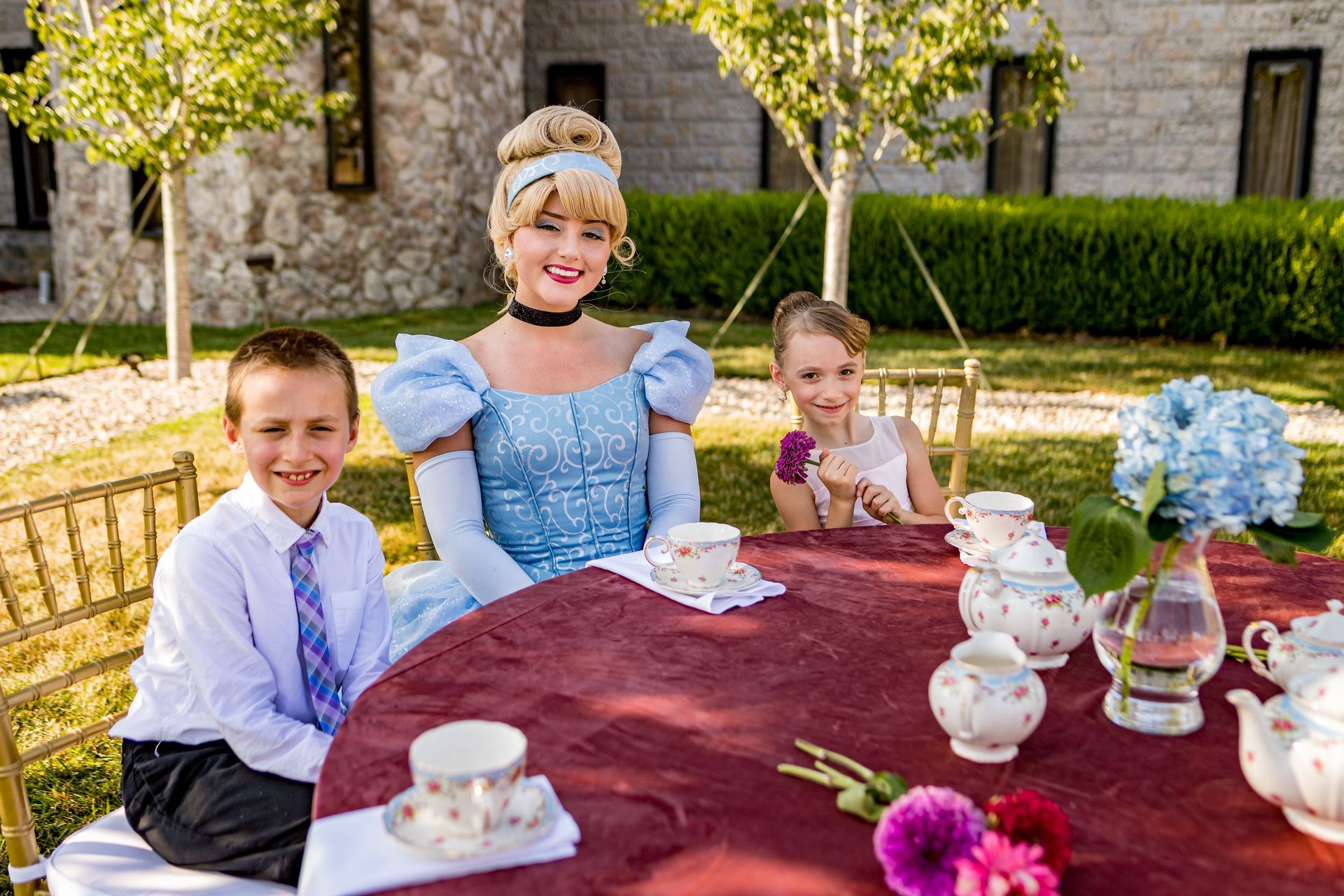 Cinderella with two children at a tea party outdoors. They sit at a red table set with tea things; blue sky.