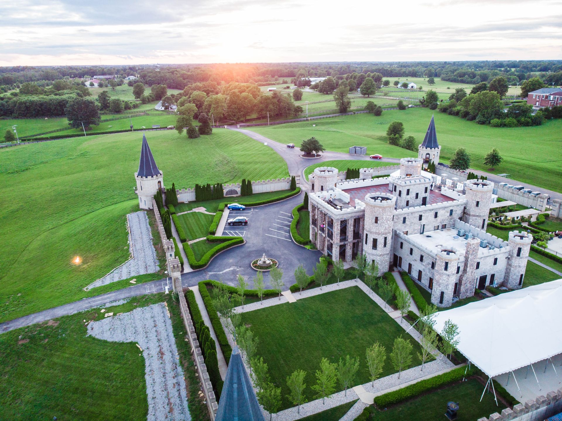 Aerial view of a light stone castle with spires, surrounded by green fields, with a sunset in the background.