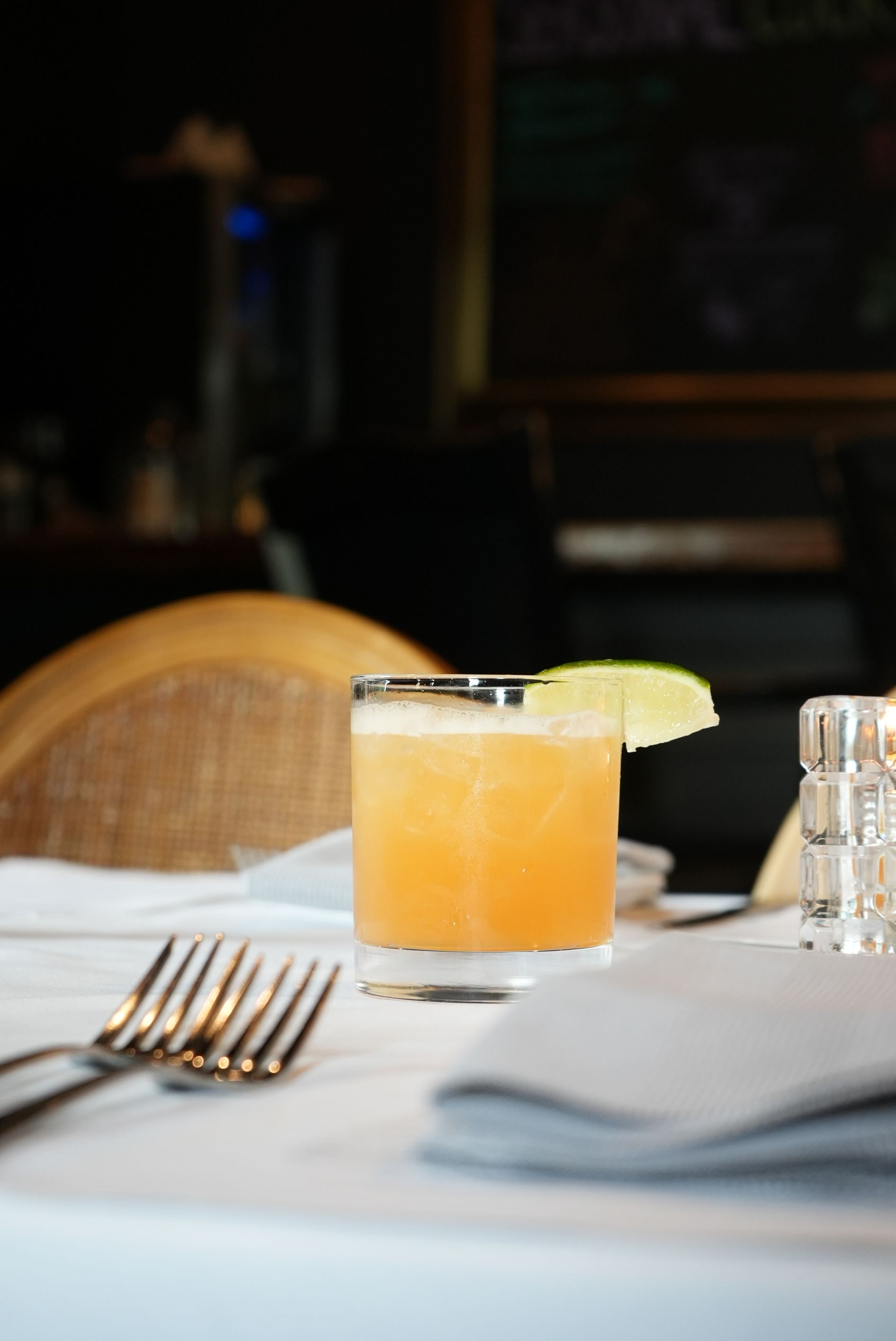 Cocktail on a table with a lime garnish, silver rimmed glass. Fork and napkin. Dark restaurant setting.