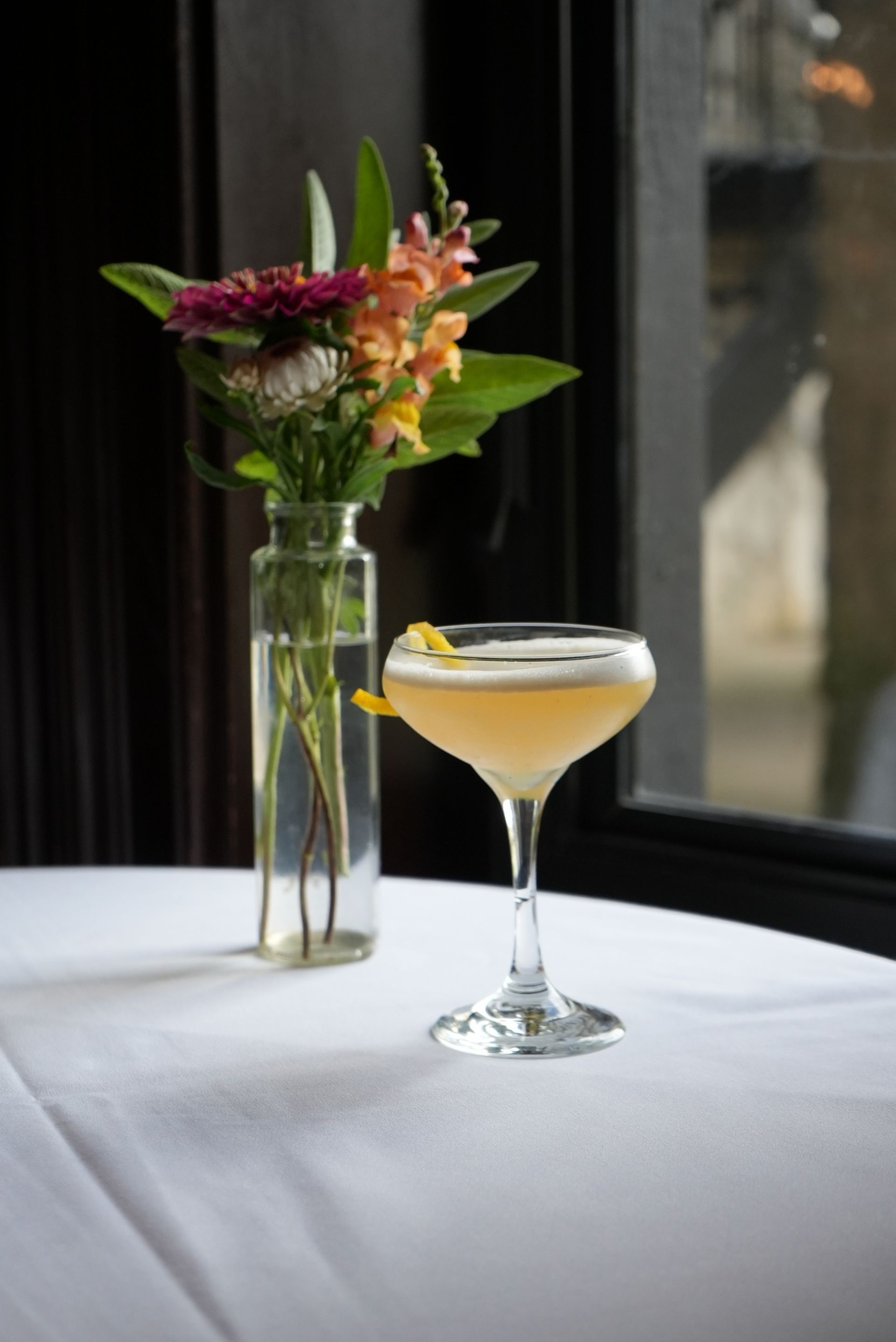 Cocktail on white tablecloth with flowers in vase next to window.