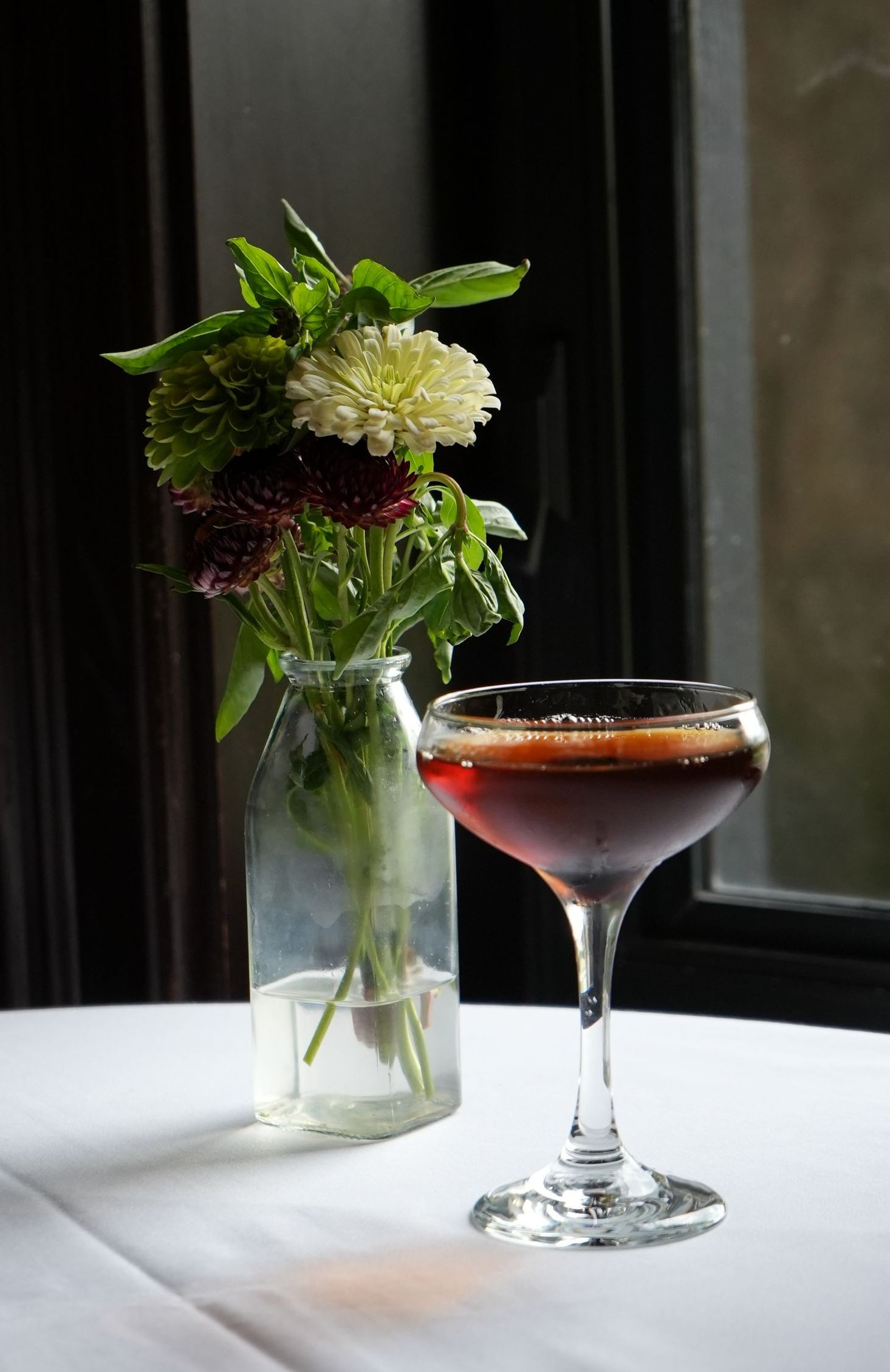 Cocktail in a coupe glass on a white tablecloth next to a small flower vase. Dark, moody aesthetic.