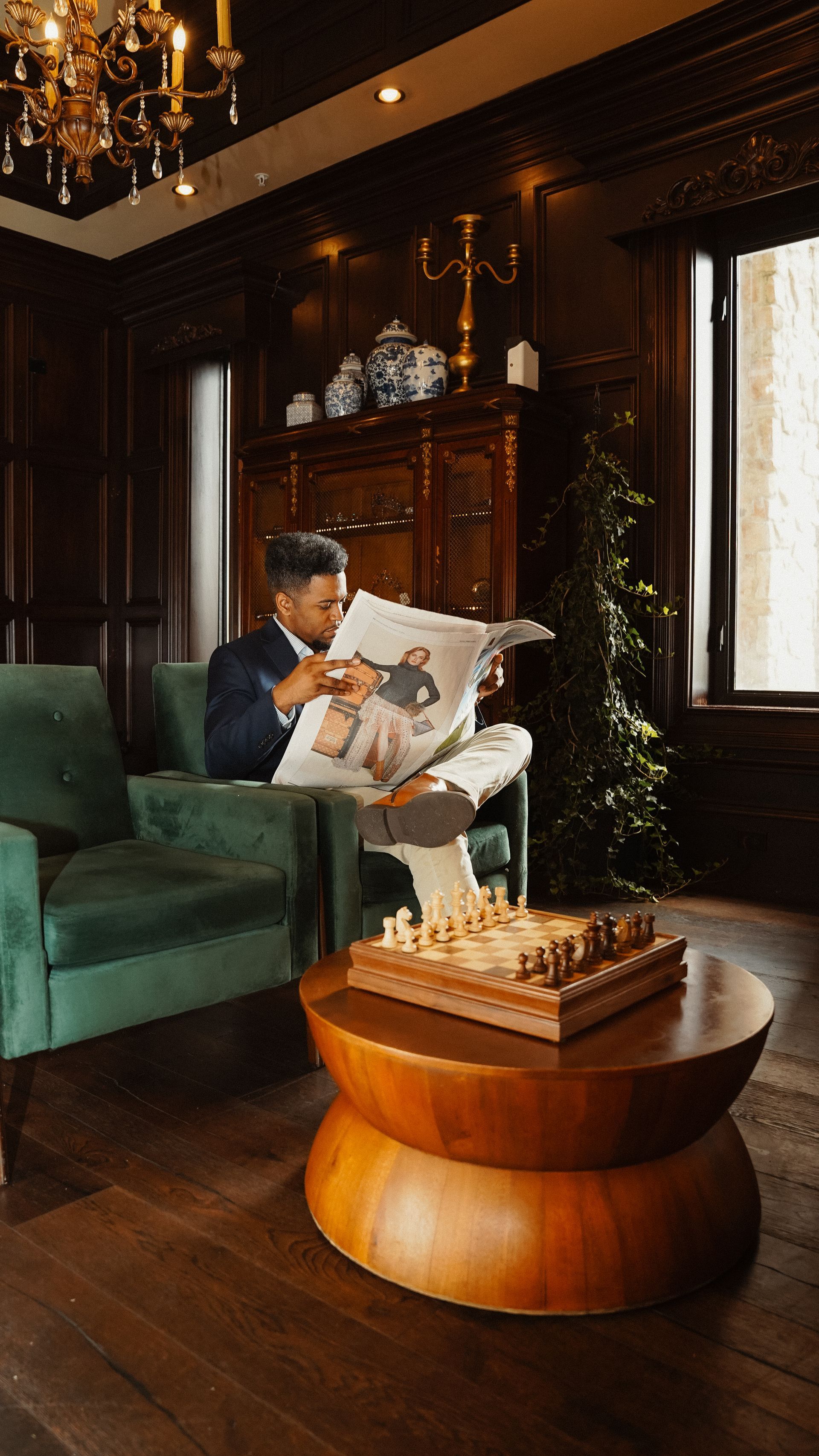 Man in suit reads newspaper in a wood-paneled room. Chessboard on a table; green velvet chair.