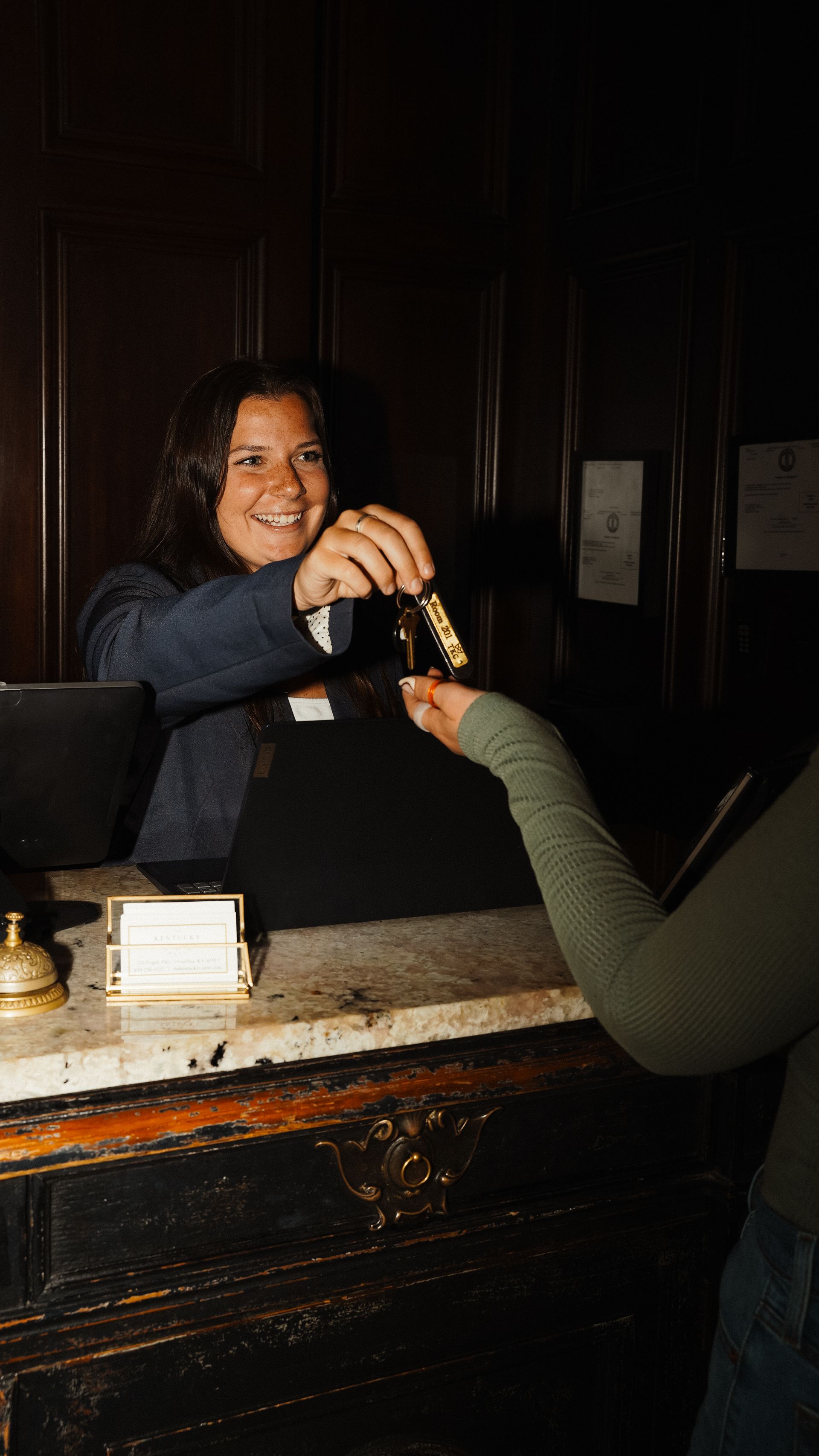 Hotel receptionist handing keys to a guest over a dark wooden desk.