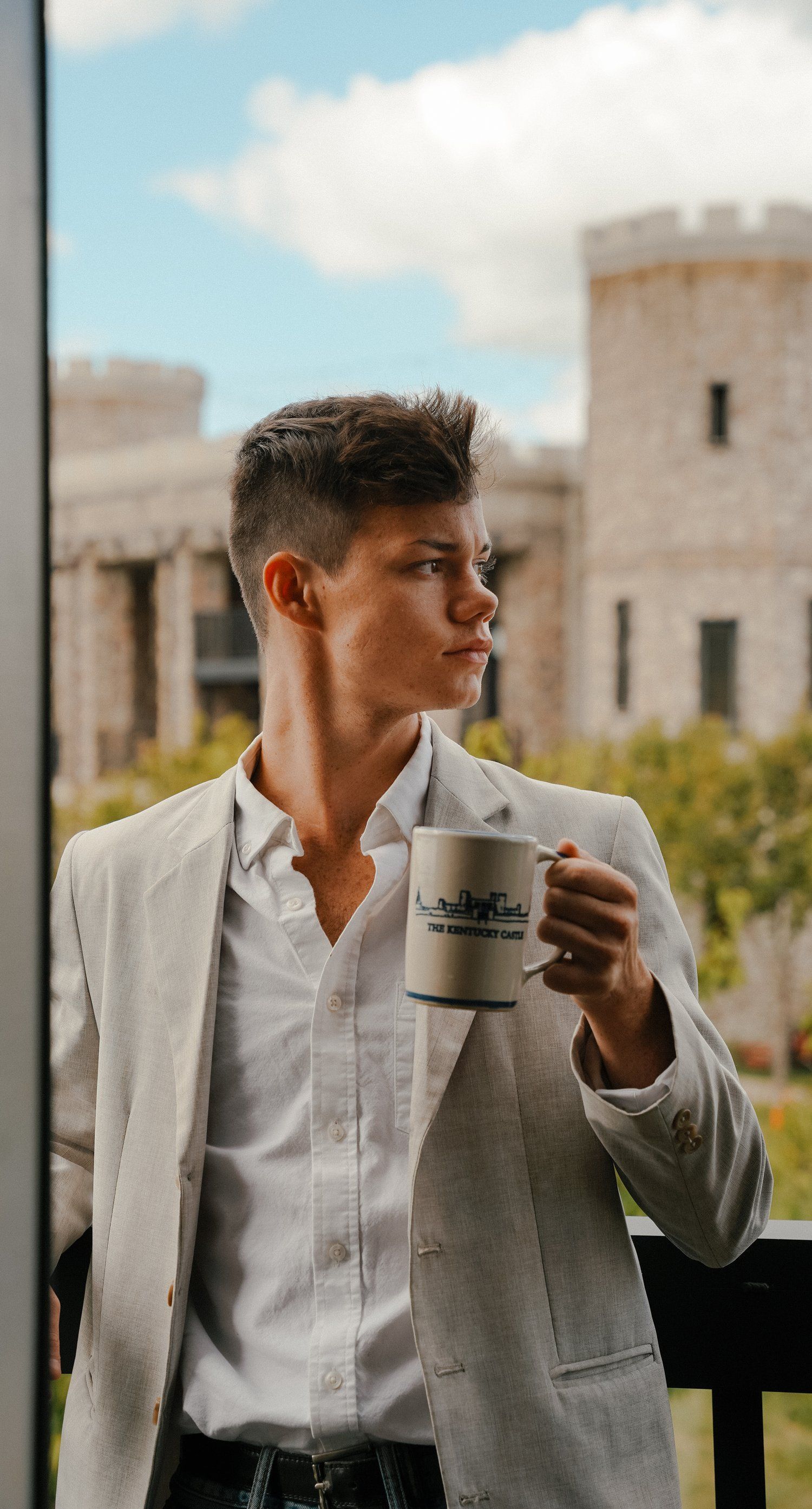 Young man in a light blazer holds a mug, looking off to the side. Castle in the background.