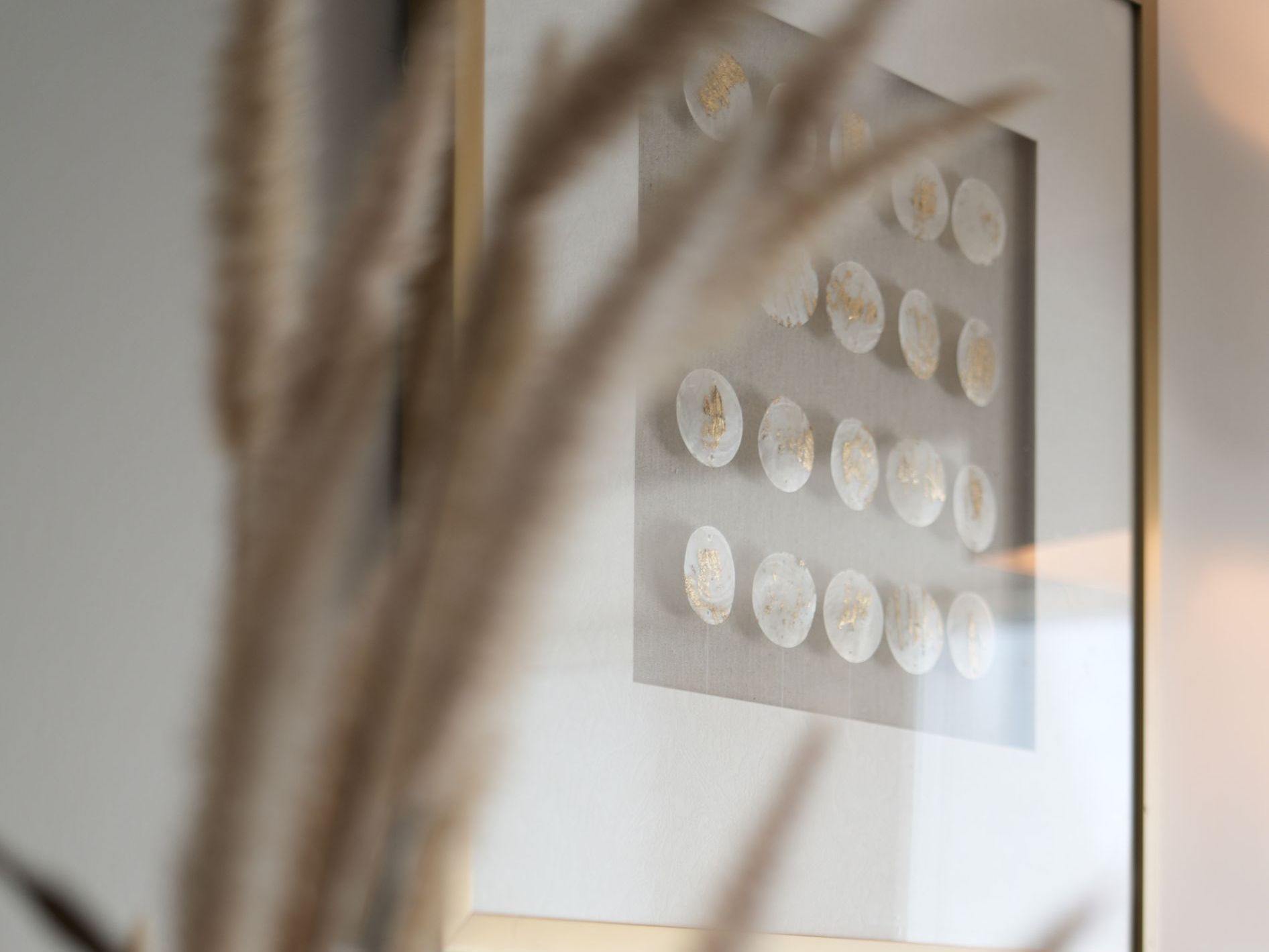 A framed artwork with several small, gold-toned circles. Foreground is blurred with tan reeds.