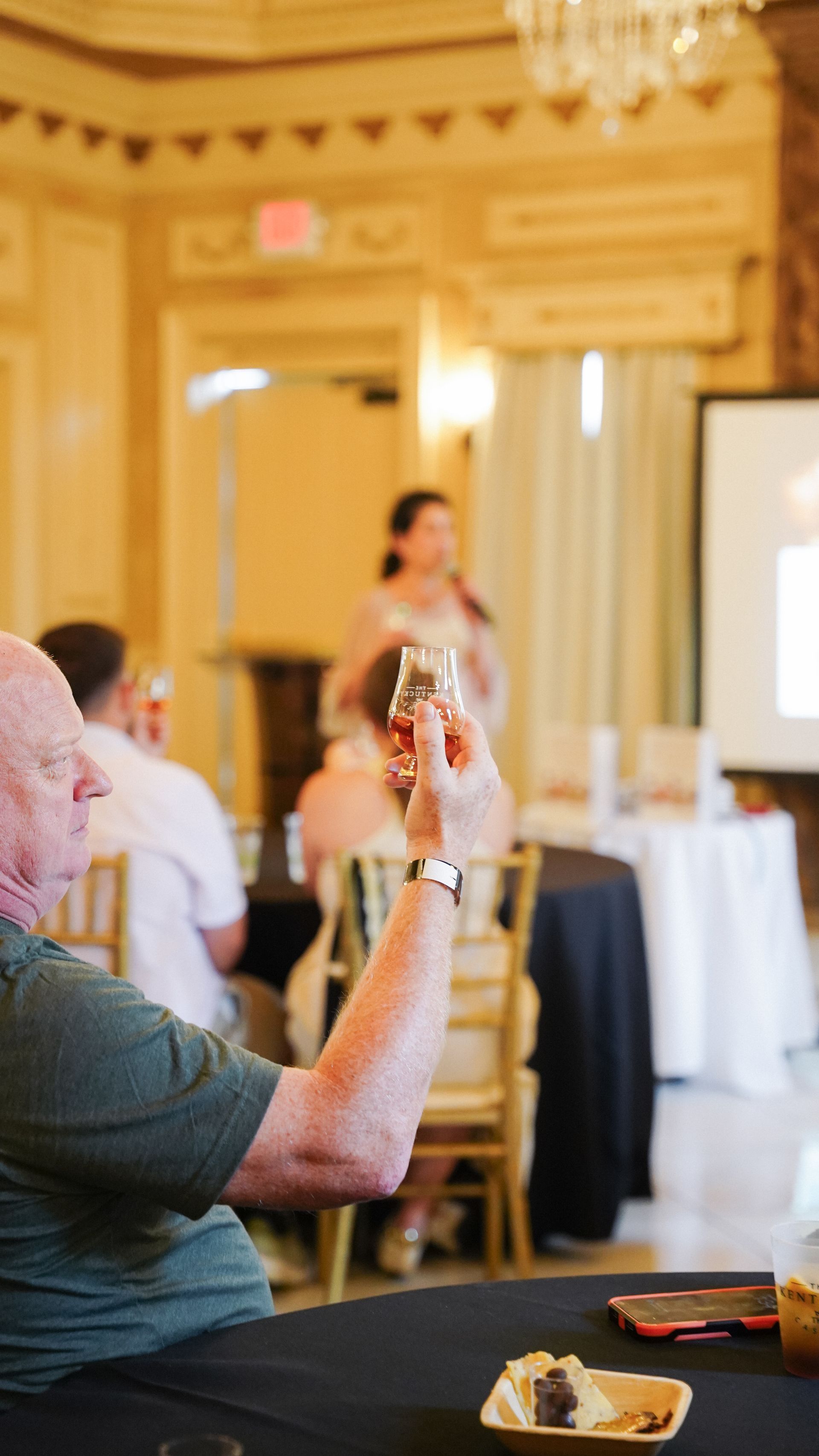 Man raises a whiskey glass in a tasting room. A presenter stands behind a table, giving a speech.