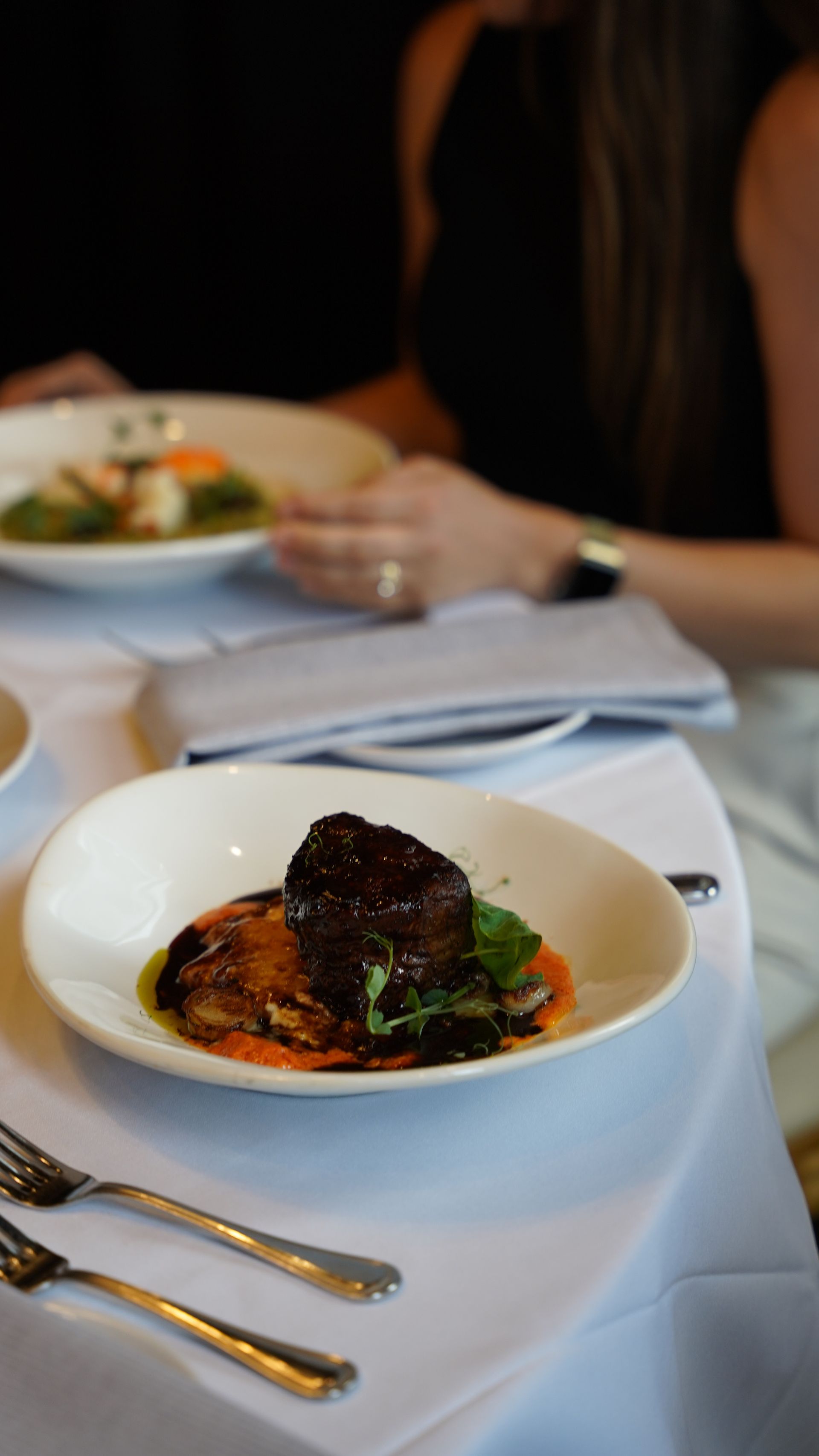 A plate with steak and vegetables on a white tablecloth. Two people are seated at the table.