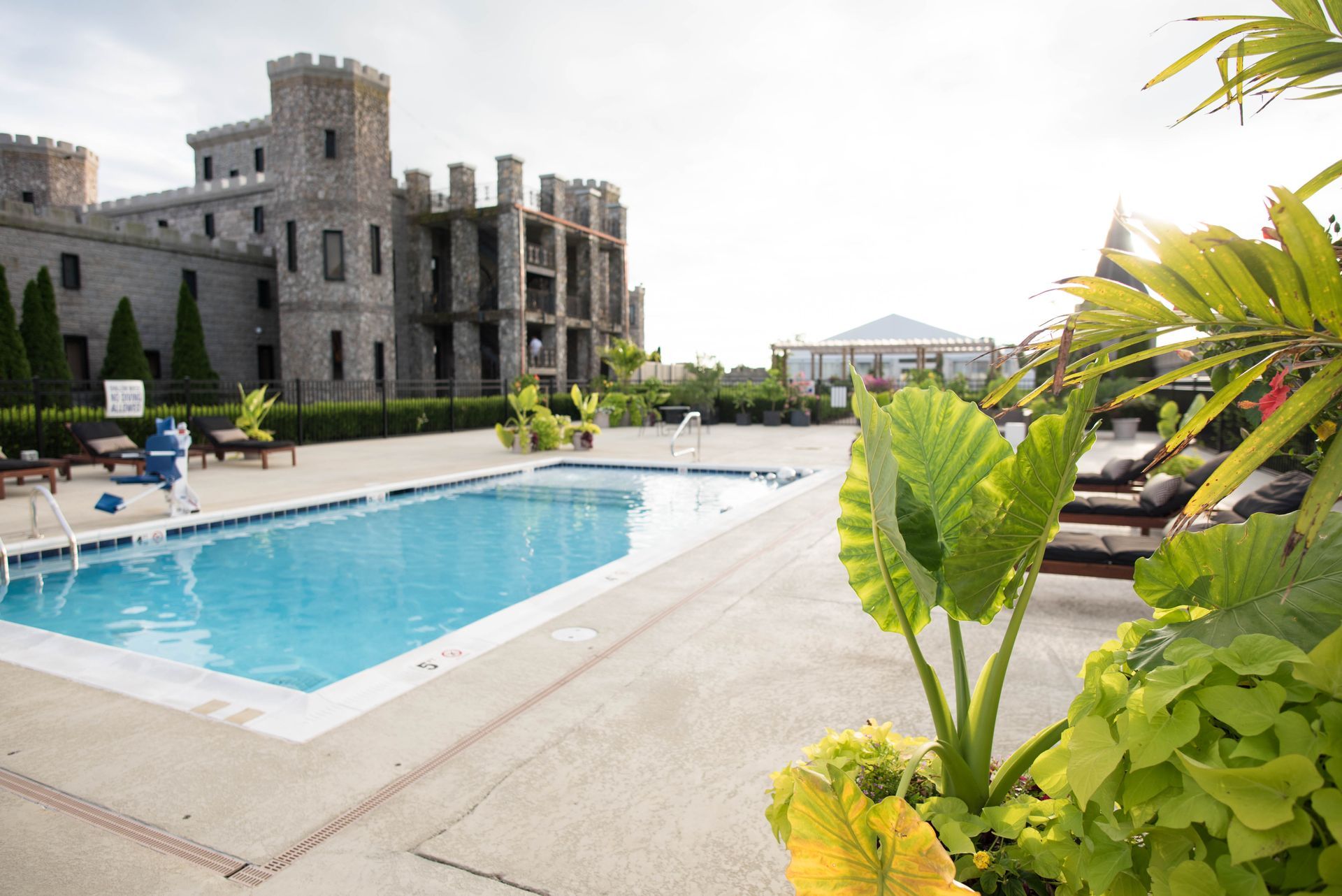 Swimming pool next to a stone castle. Lounge chairs and lush greenery surround the pool on a sunny day.