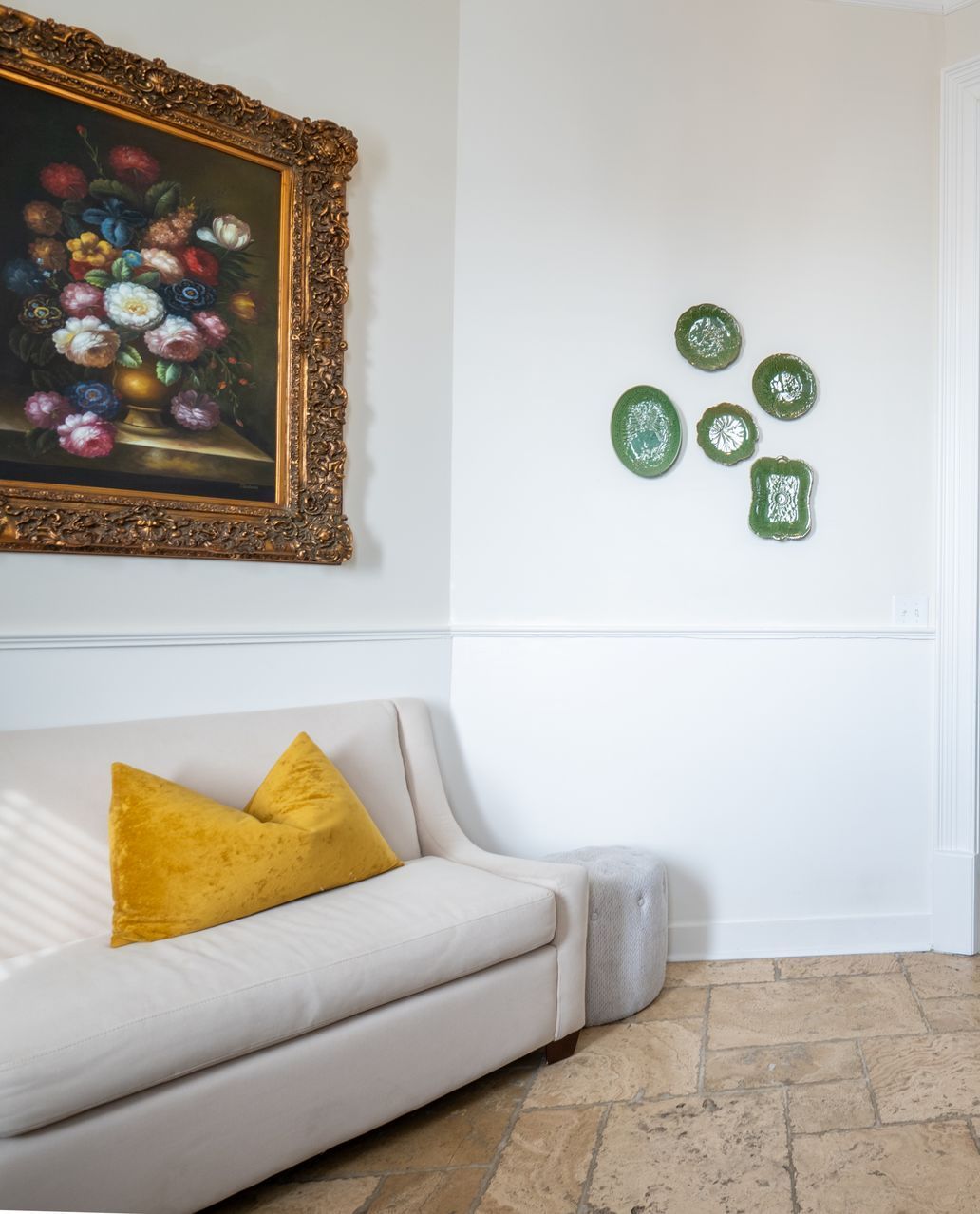 A chaise lounge with yellow pillow beside floral painting and green plates on white walls.