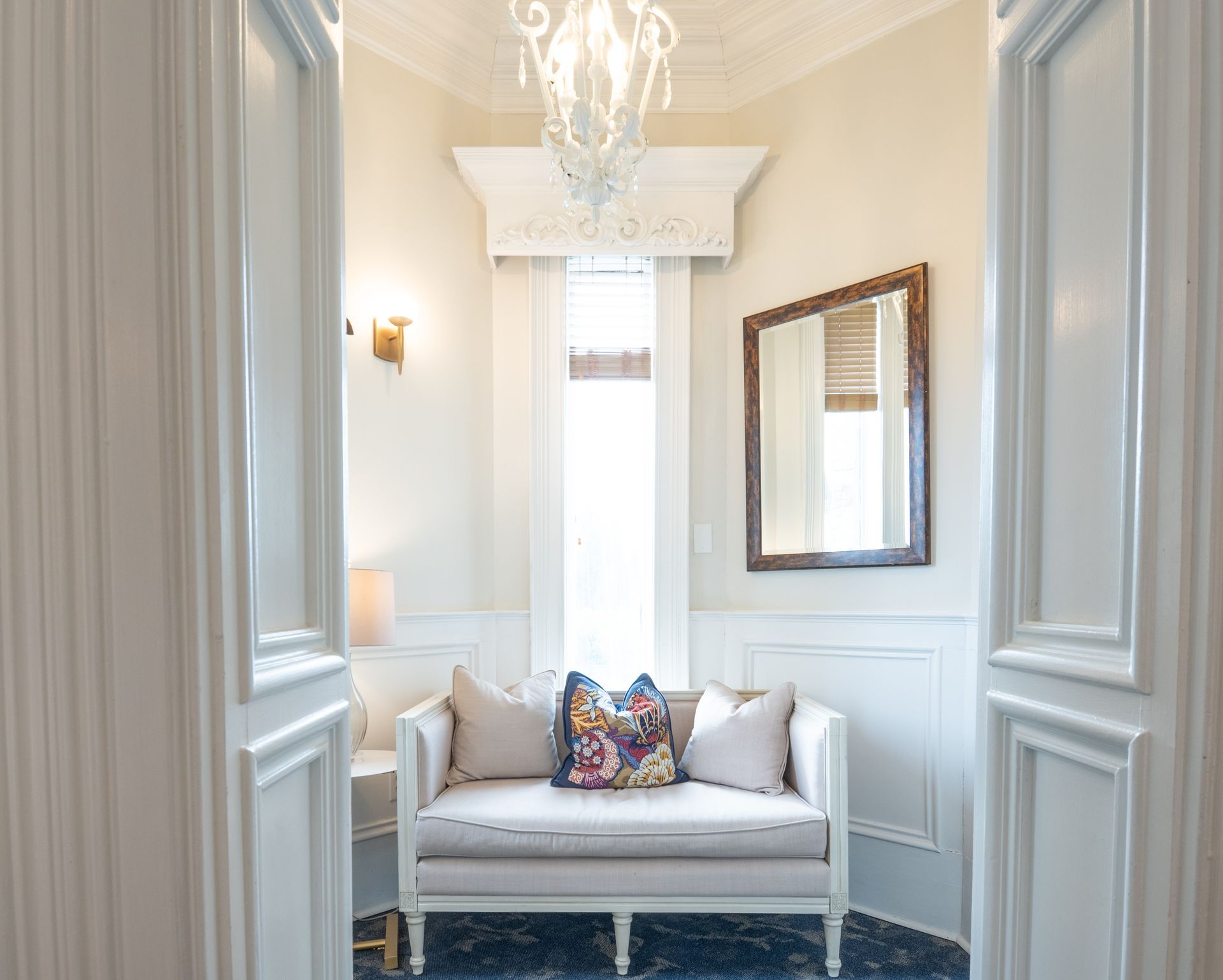White-walled entryway with a cream-colored loveseat, mirror, and ornate chandelier.