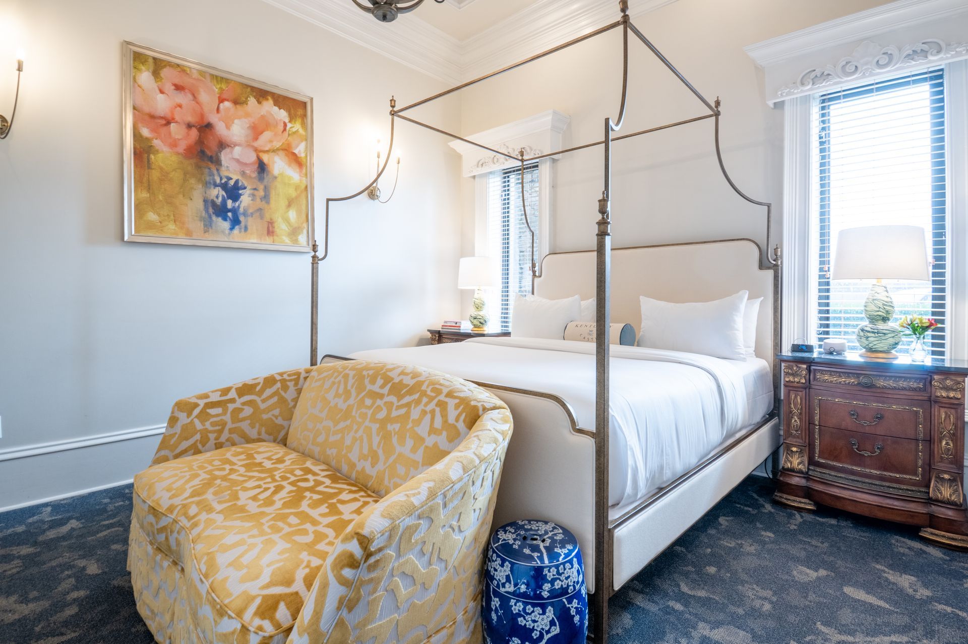 Bedroom with four-poster bed, ornate chair, and floral art, featuring gold and blue accents.