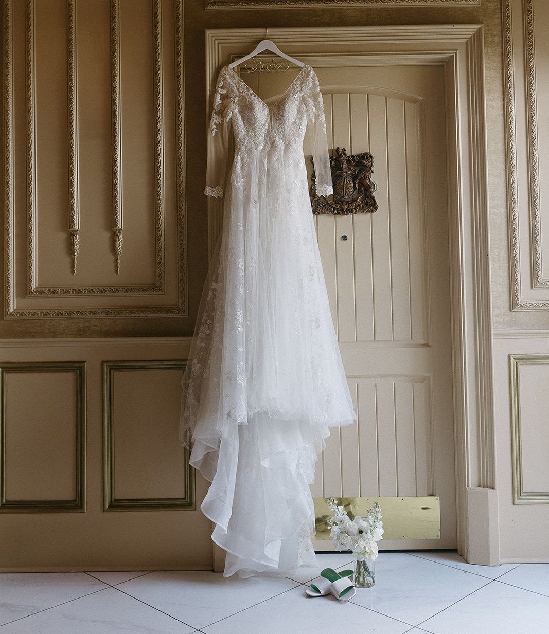 Wedding dress hanging on a door; white, long-sleeved, flowing, and a short train. Flowers and shoes below.