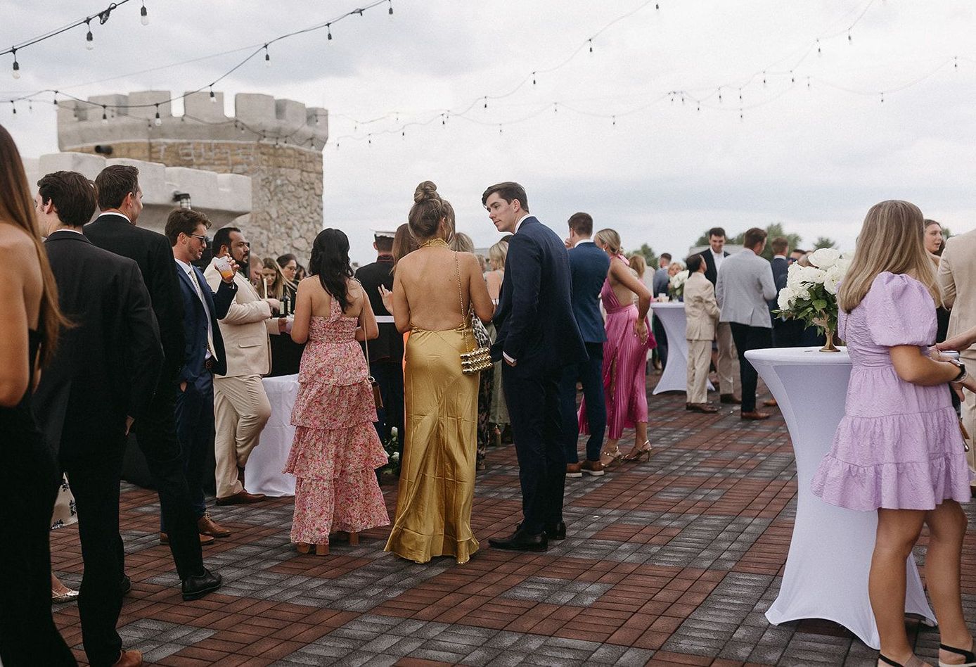 People in formal attire at an outdoor event; a castle-like structure is in the background.