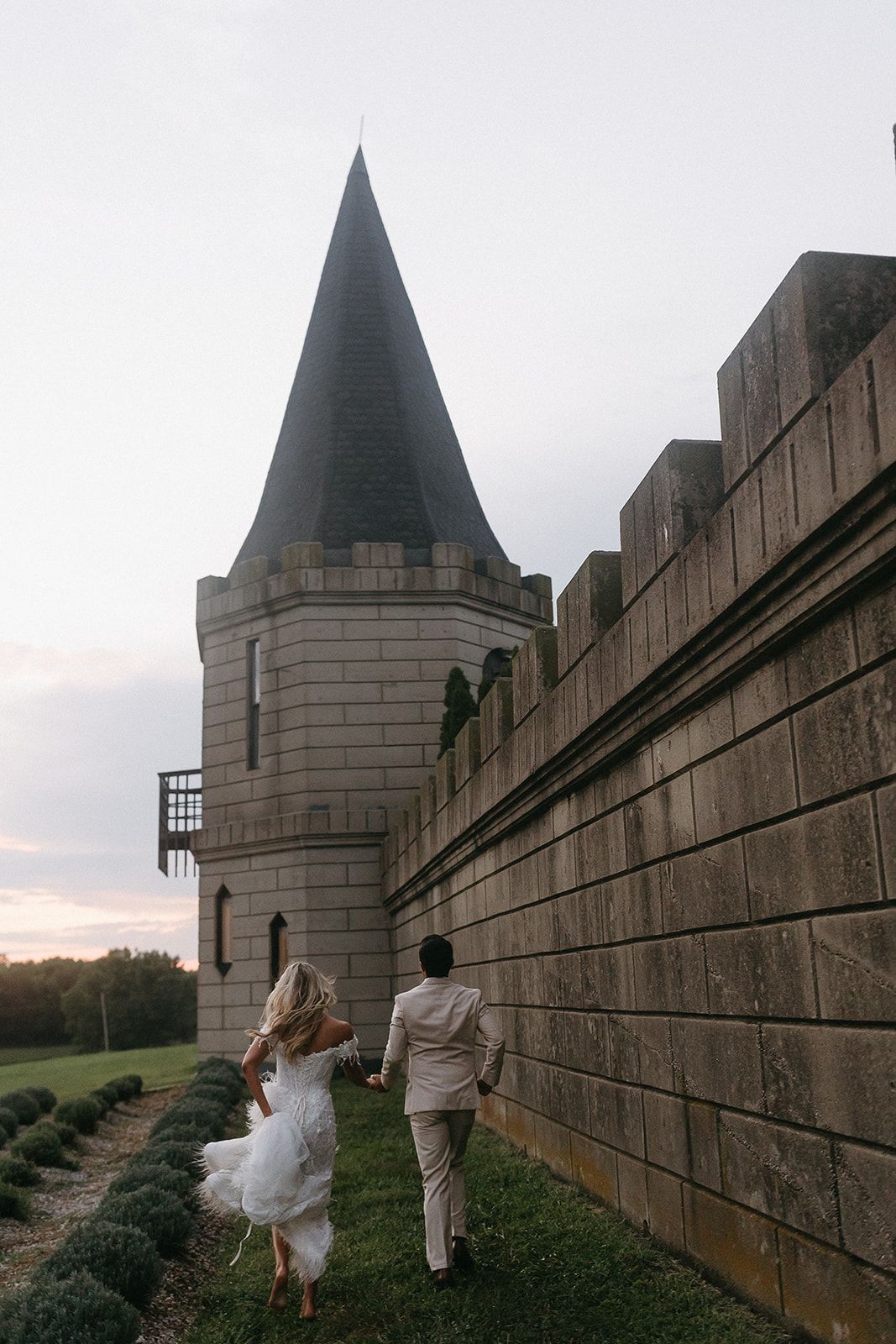 Bride and groom run, holding hands, along a castle wall in a field at dusk.