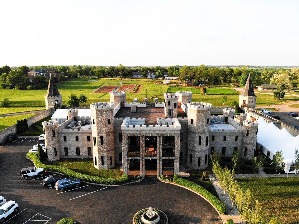 Stone castle with turrets, central open area, circular driveway, and cars parked outside in a green field.