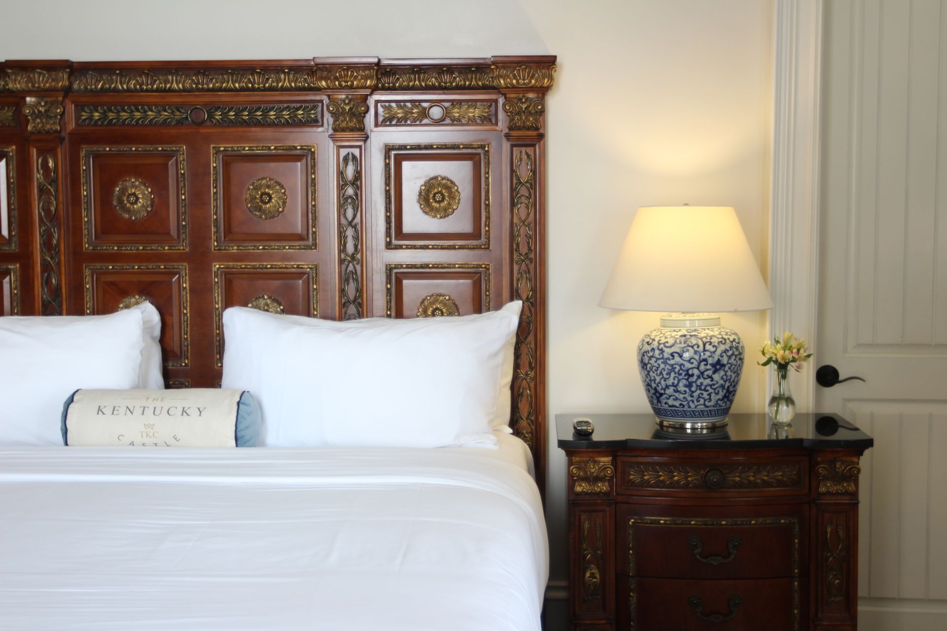 Bedroom with a dark ornate headboard, white bedding, a blue and white lamp, and a bedside table.