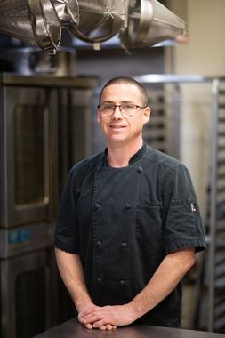 Chef in black uniform, glasses, smiling in a kitchen, ovens in background.