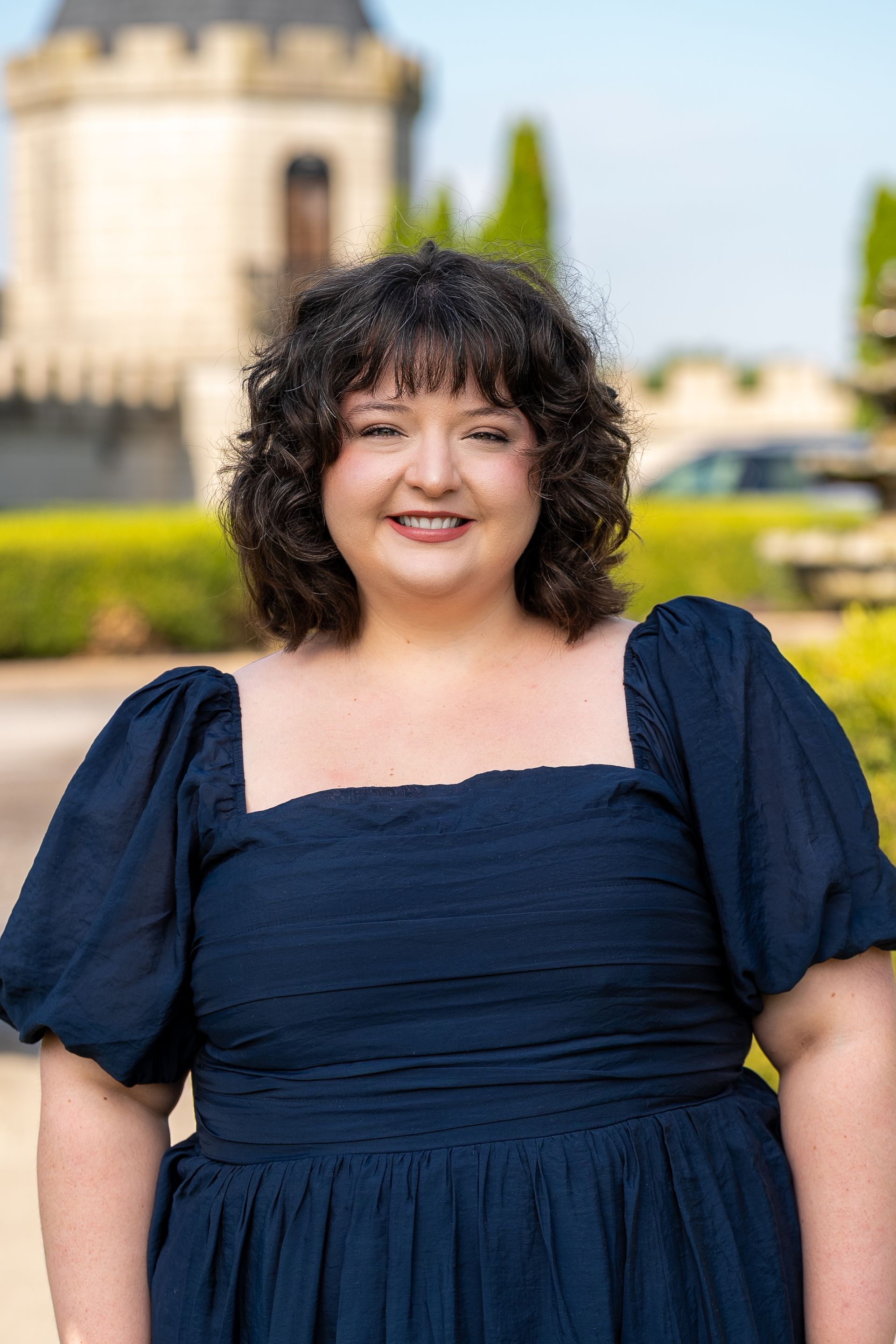 Woman with long brown curly hair in blue polka dot dress smiles, leaning on a marble counter.