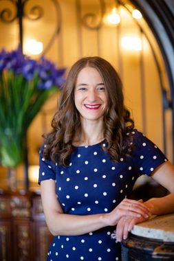 Woman with long brown curly hair in blue polka dot dress smiles, leaning on a marble counter.