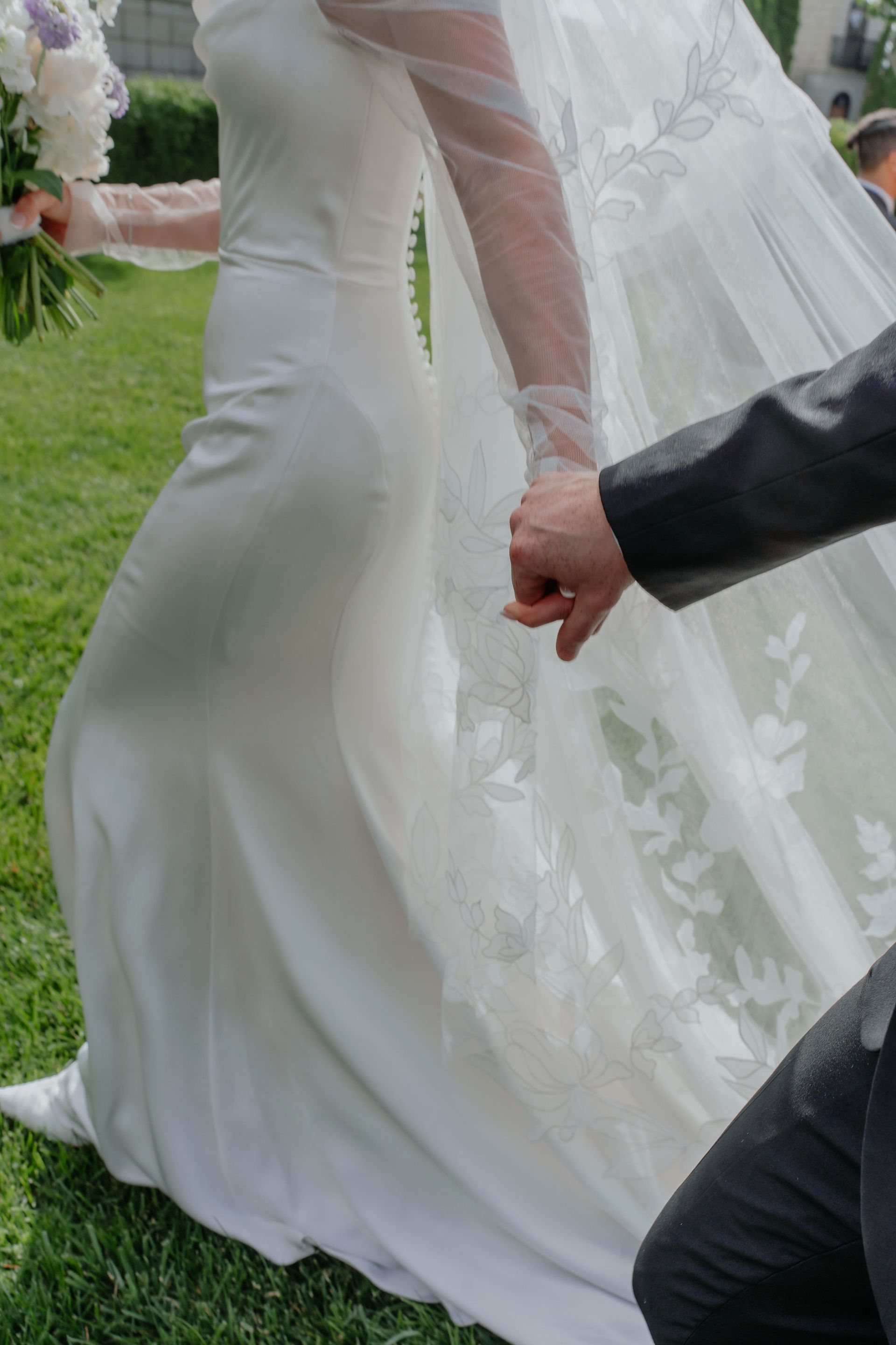 Bride in a white satin dress and veil holds hands with a person in a black suit, outdoors.