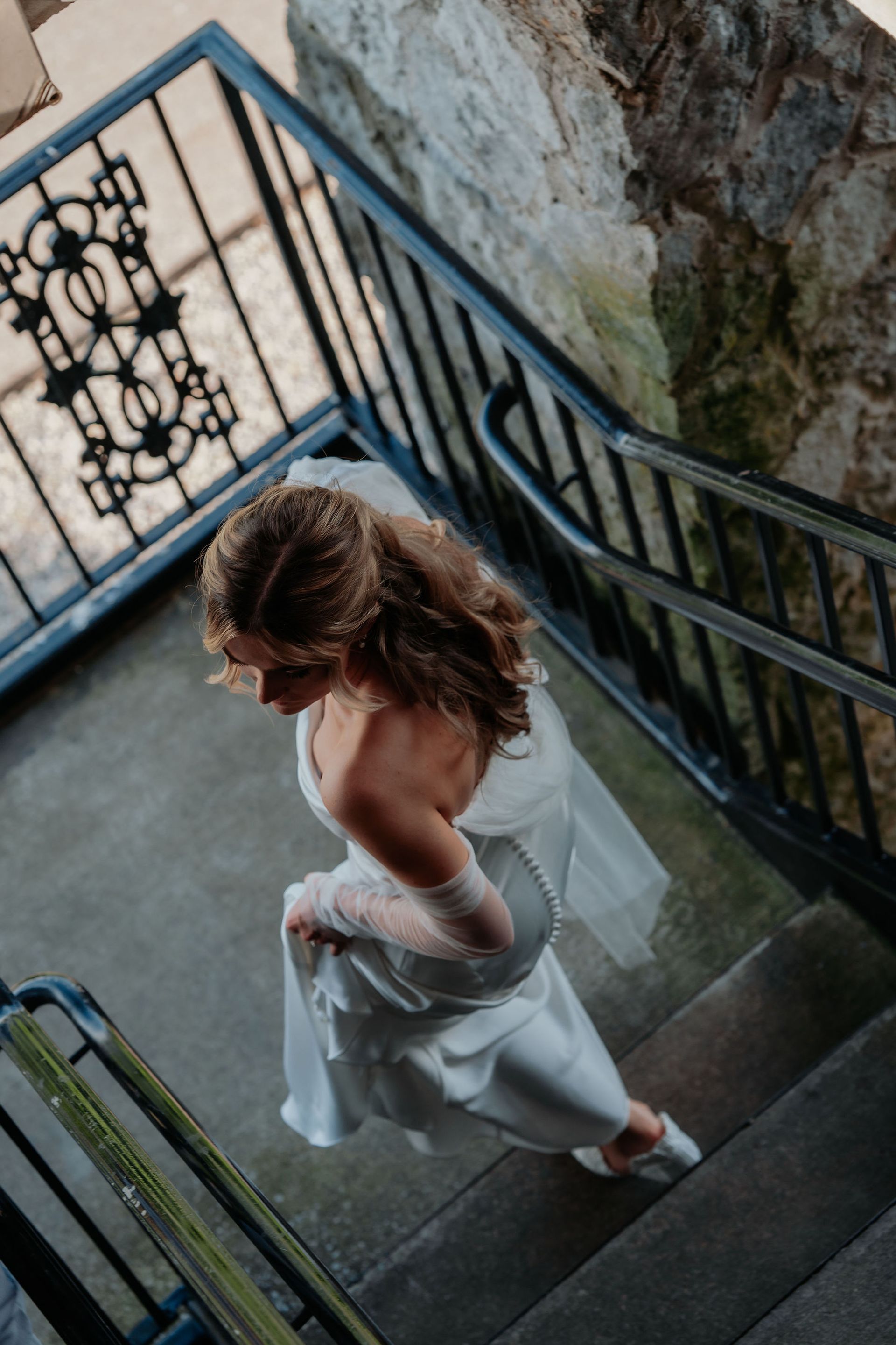 Bride in white dress descends stone stairs, with ornate black railing.