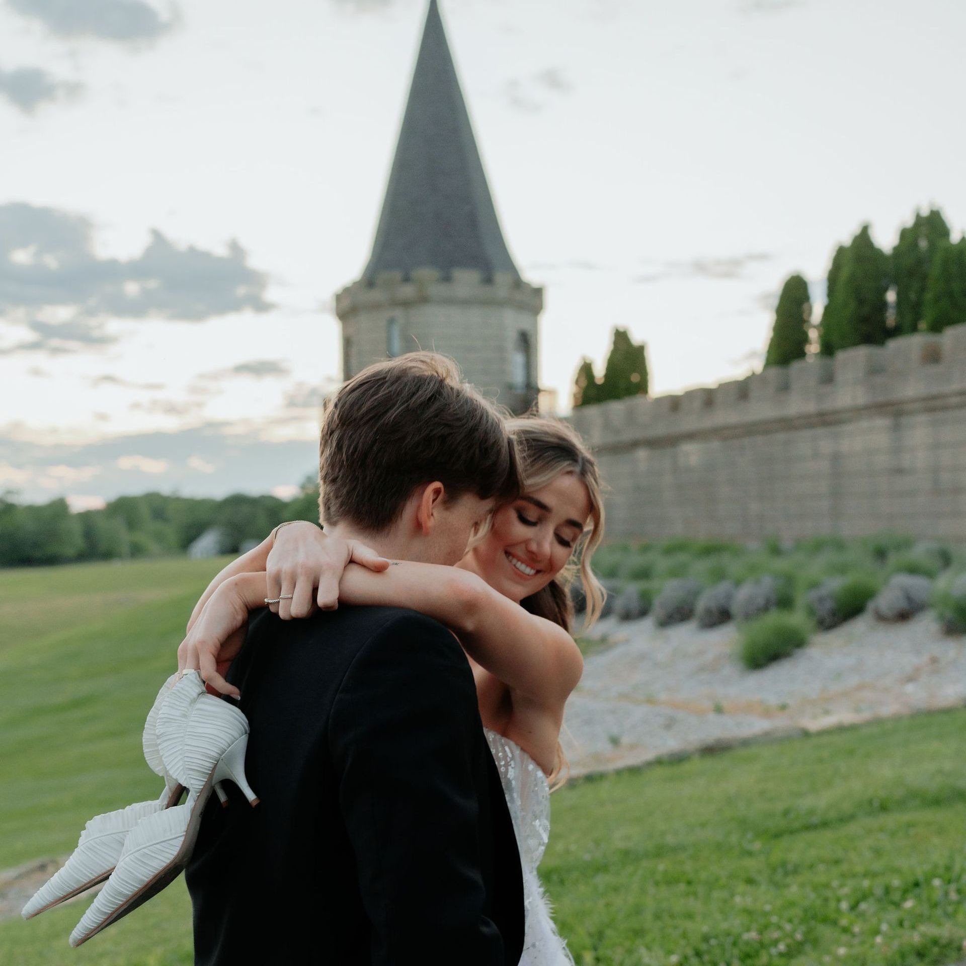 Newlyweds embrace, smiling, outside castle, bride holding heels, against a grassy landscape.