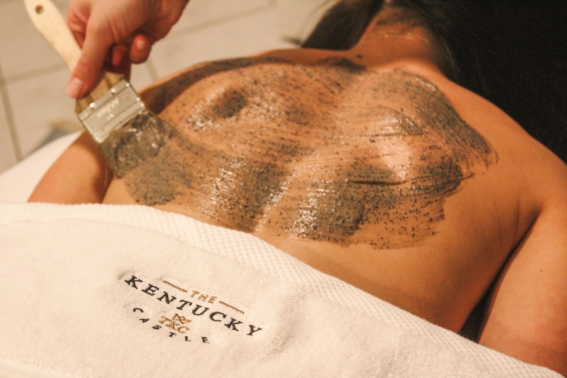 Person receiving a back treatment at a spa. A hand applies a dark scrub with a brush to the back. White towel.