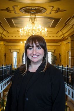 Woman smiling in an ornate, gold-toned hallway, with chandelier above. She wears a black jacket.