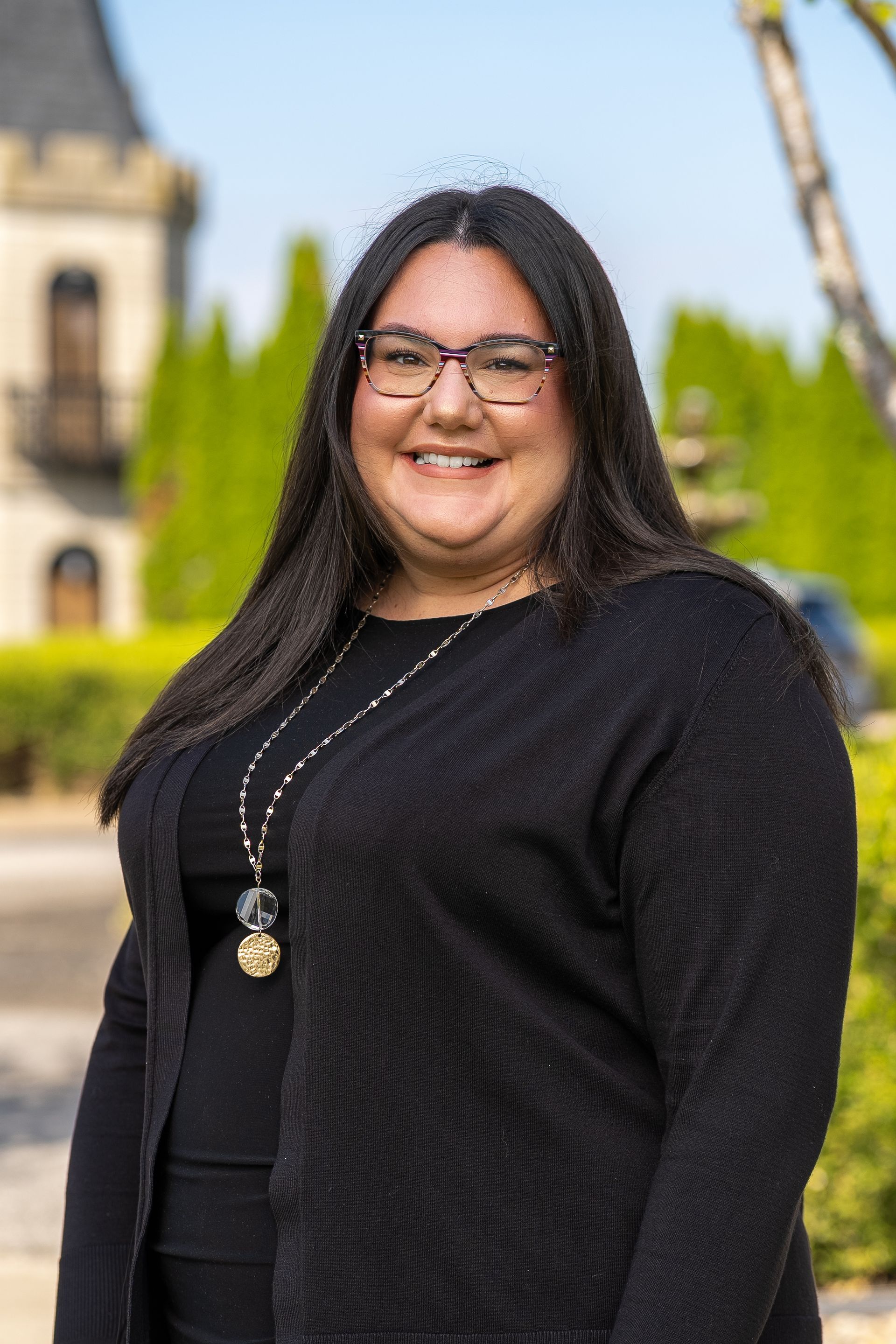Chef in black uniform, glasses, smiling in a kitchen, ovens in background.