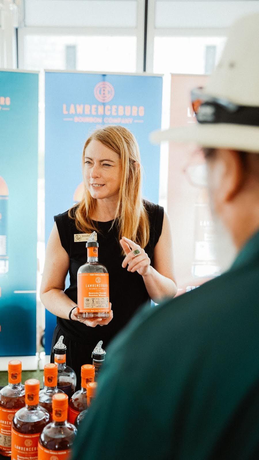 Woman presenting liquor, holding a bottle, speaking to a person. Bottles on a table. Event setting with banners.