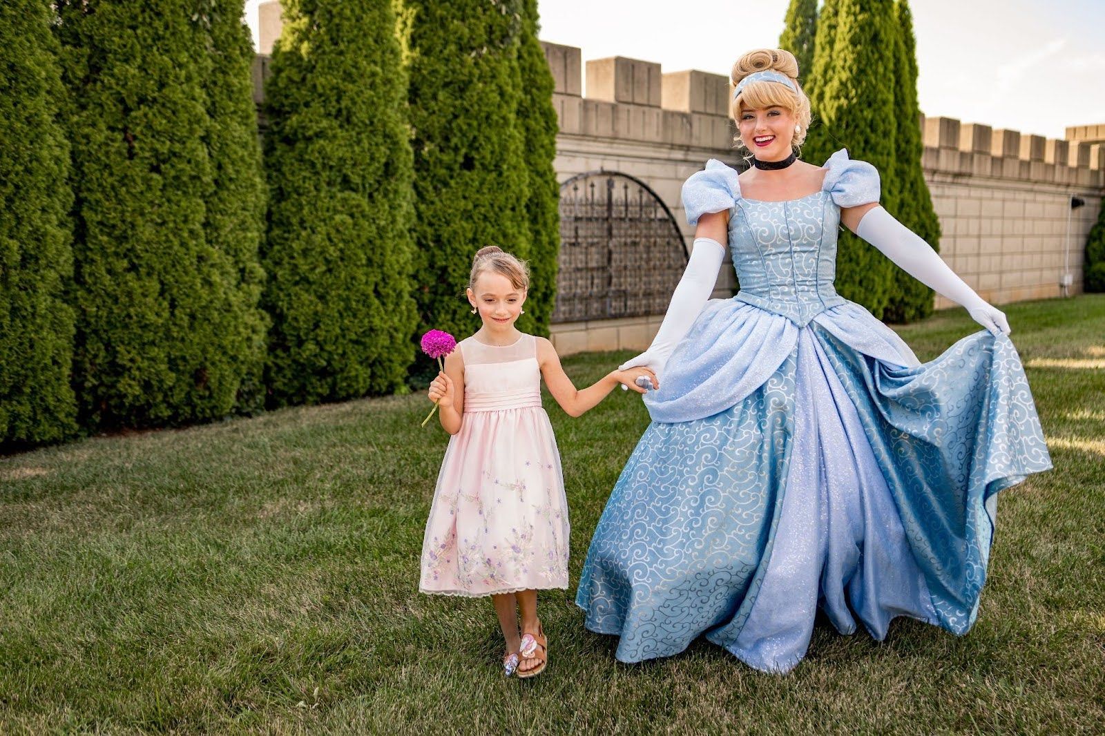 Cinderella in blue gown holding hands with a girl in pink dress on lawn in front of a castle wall.