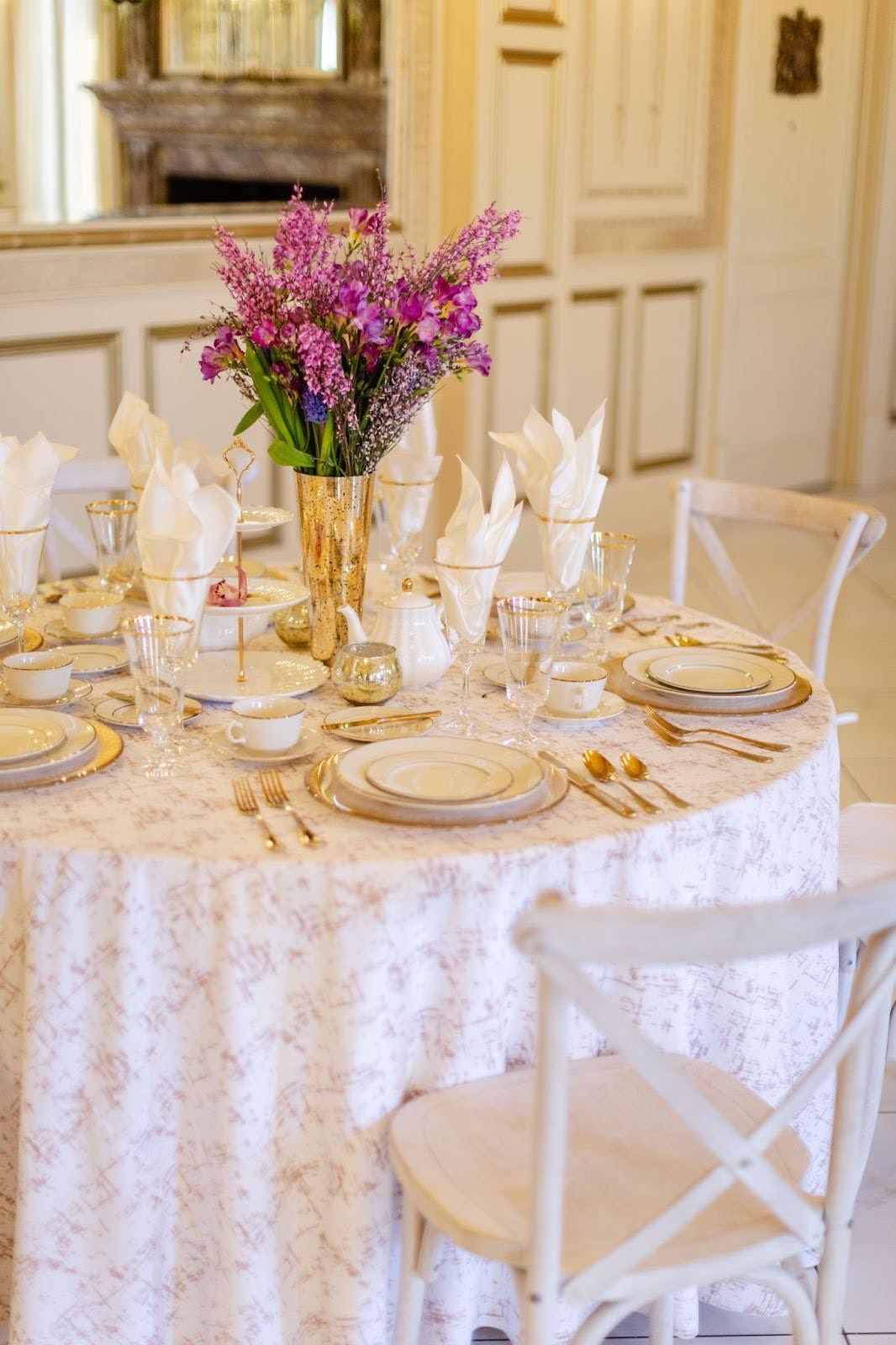 Round table set for a meal with gold accents, purple flowers in vase, and white chairs.