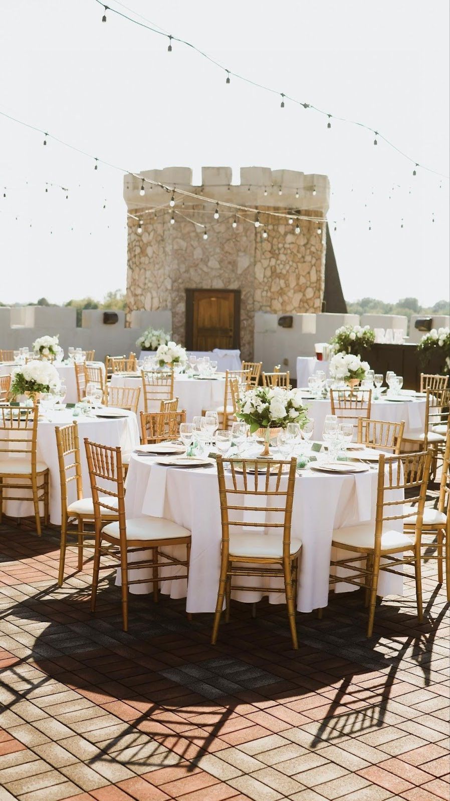 Wedding reception on a rooftop with tables, chairs, flowers, and a castle-like tower. Gold and white decor.