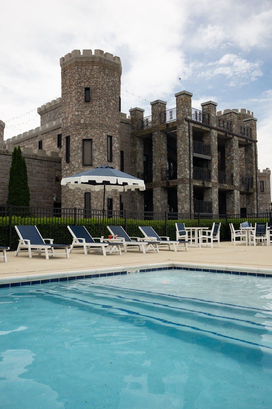Castle hotel with a pool, blue lounge chairs, and a striped umbrella. Cloudy sky.