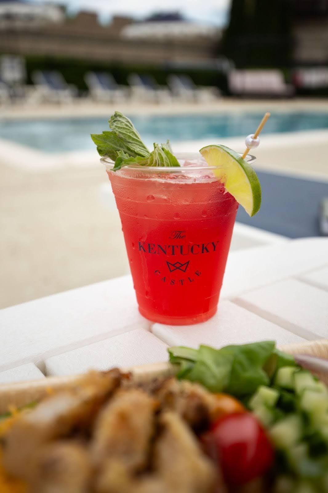 Red cocktail in plastic cup with lime and mint by a pool.
