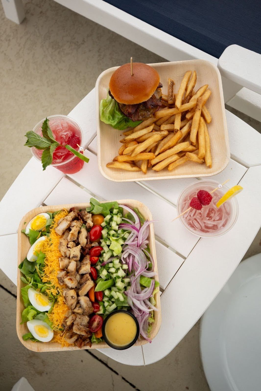 Burger and fries, chicken salad, and two drinks on a white table by a pool.