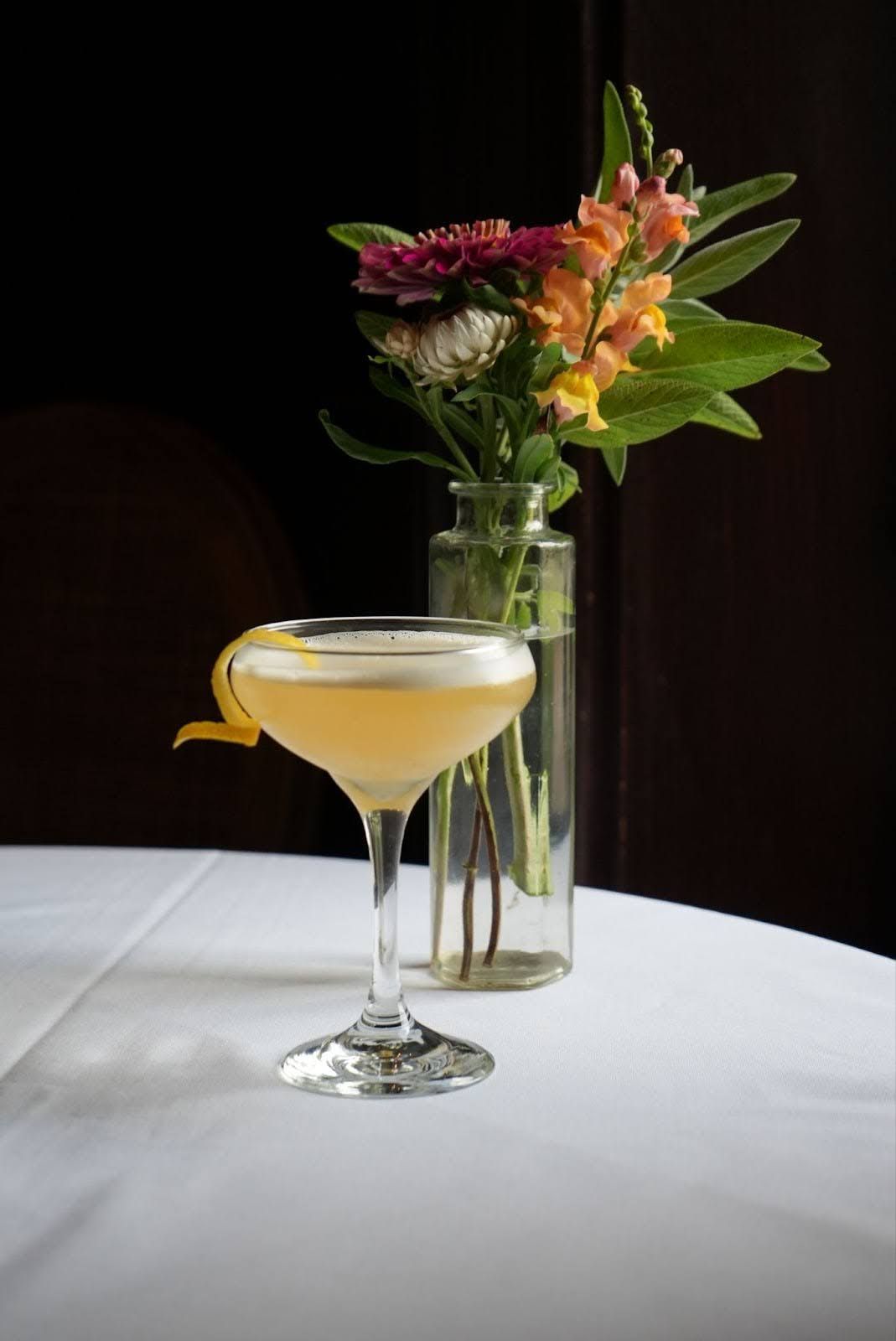 Cocktail in a stemmed glass with a lemon twist, next to a vase of colorful flowers, on a white tablecloth.